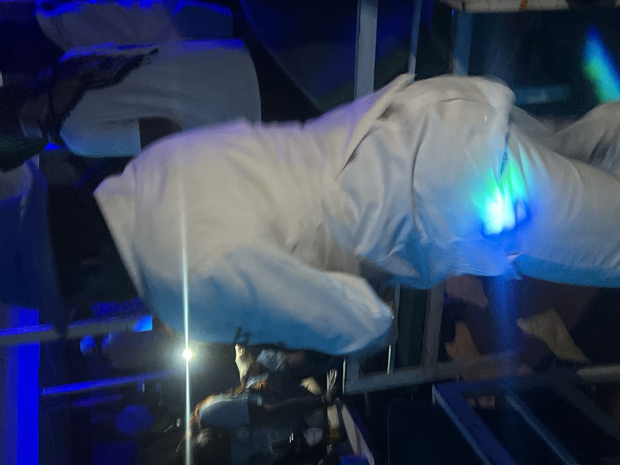 The image depicts a dimly lit indoor scene, likely a club or social gathering, with predominantly blue and green lighting. A person in the foreground is wearing a white outfit, including pants and a jacket, and a hat. Their back is to the camera, and they are illuminated by a bright green and blue light emanating from their lower back area. Other individuals are visible in the background, also in low light conditions, suggesting a social event. There are hints of furniture like chairs and tables, typical of a nightlife venue. The scene appears to be taking place in Durumi, Nigeria, given the prompt's specification.