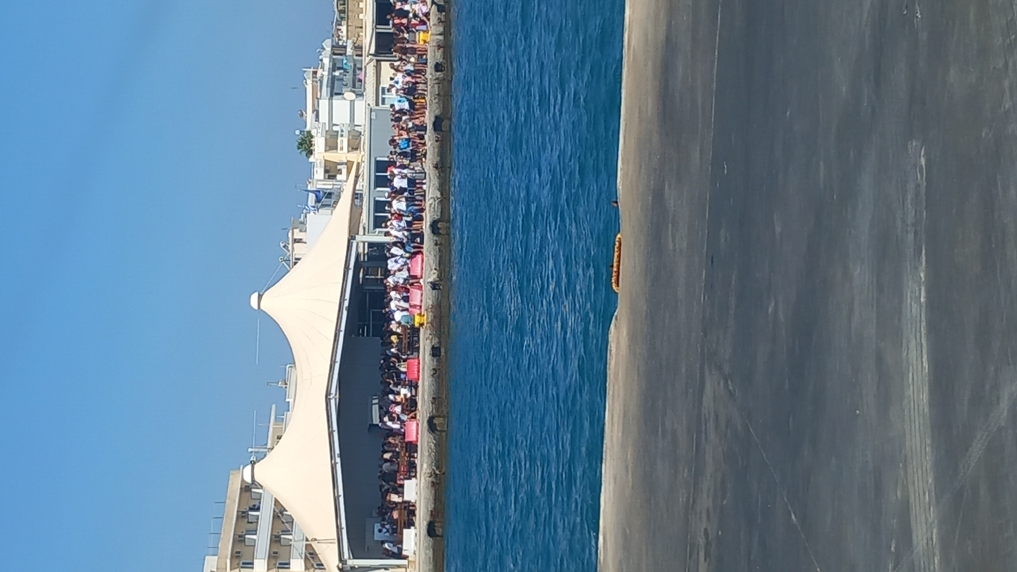 In Kos, Greece, a crowd of people are gathered on a pier. The pier is lined with benches and a large tent-like structure. The sea is a vibrant blue, and buildings can be seen in the background. The sky is clear and blue.