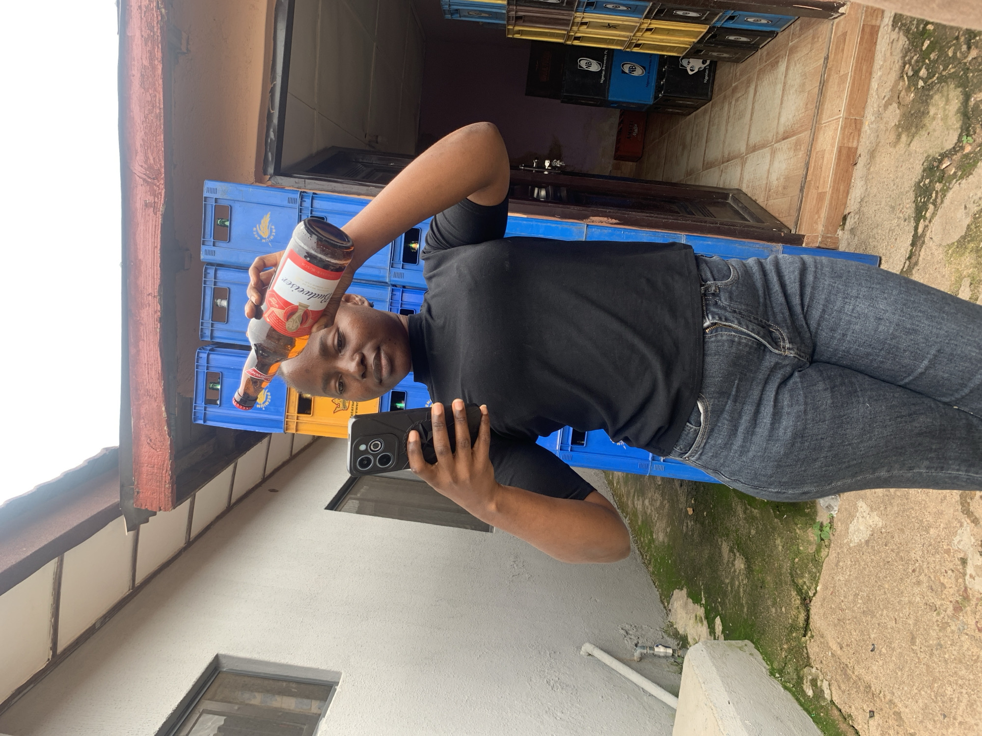 The image shows a person holding a bottle of Budweiser and a smartphone. The person is wearing a black t-shirt and dark jeans. They appear to be taking a selfie with the smartphone.
In the background, there are stacks of blue and yellow plastic crates, some of which have labels that are partially visible. One visible label appears to read "Nigerian Breweries Plc" and another "Amstel Malta". The setting appears to be an outdoor area, possibly a storage yard or a shop, with a concrete floor and some mossy patches. There is also a building with a white wall and a window visible to the left.
The lighting suggests it might be daytime, but the sky is overcast. The overall mood is casual. The visible text on the bottle is "Budweiser".