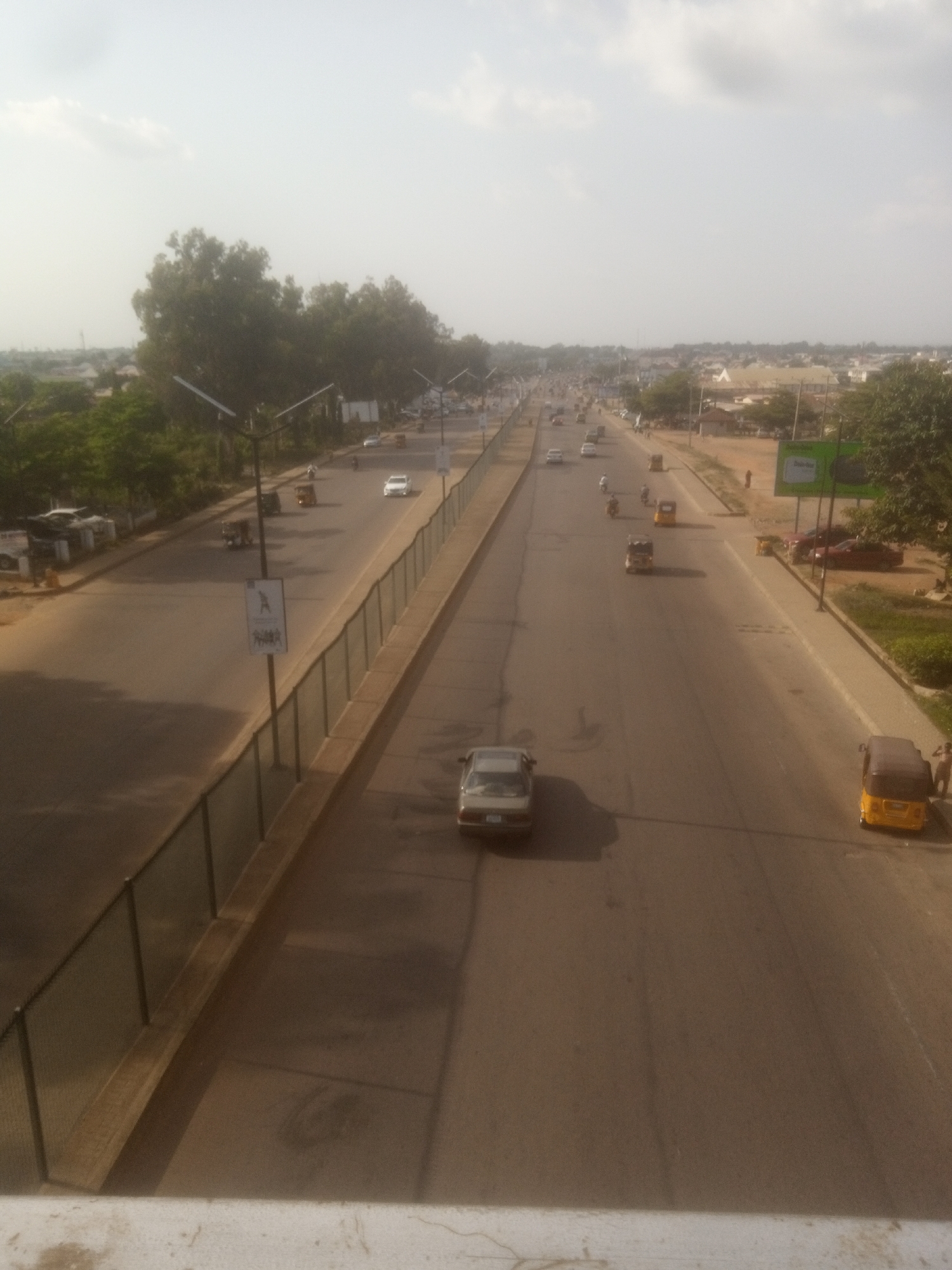 This image depicts a highway scene in Unguwan Dosa, Nigeria, characterized by a wide, paved road with multiple lanes. Several vehicles, including cars and tricycles (popularly known as 'Keke Napep'), are visible on the road, suggesting moderate traffic flow. Trees line the sides of the highway, with some buildings and a signpost also visible in the background. The sky is bright and appears to be overcast or hazy, indicating daytime. The overall impression is that of a bustling urban or peri-urban environment.