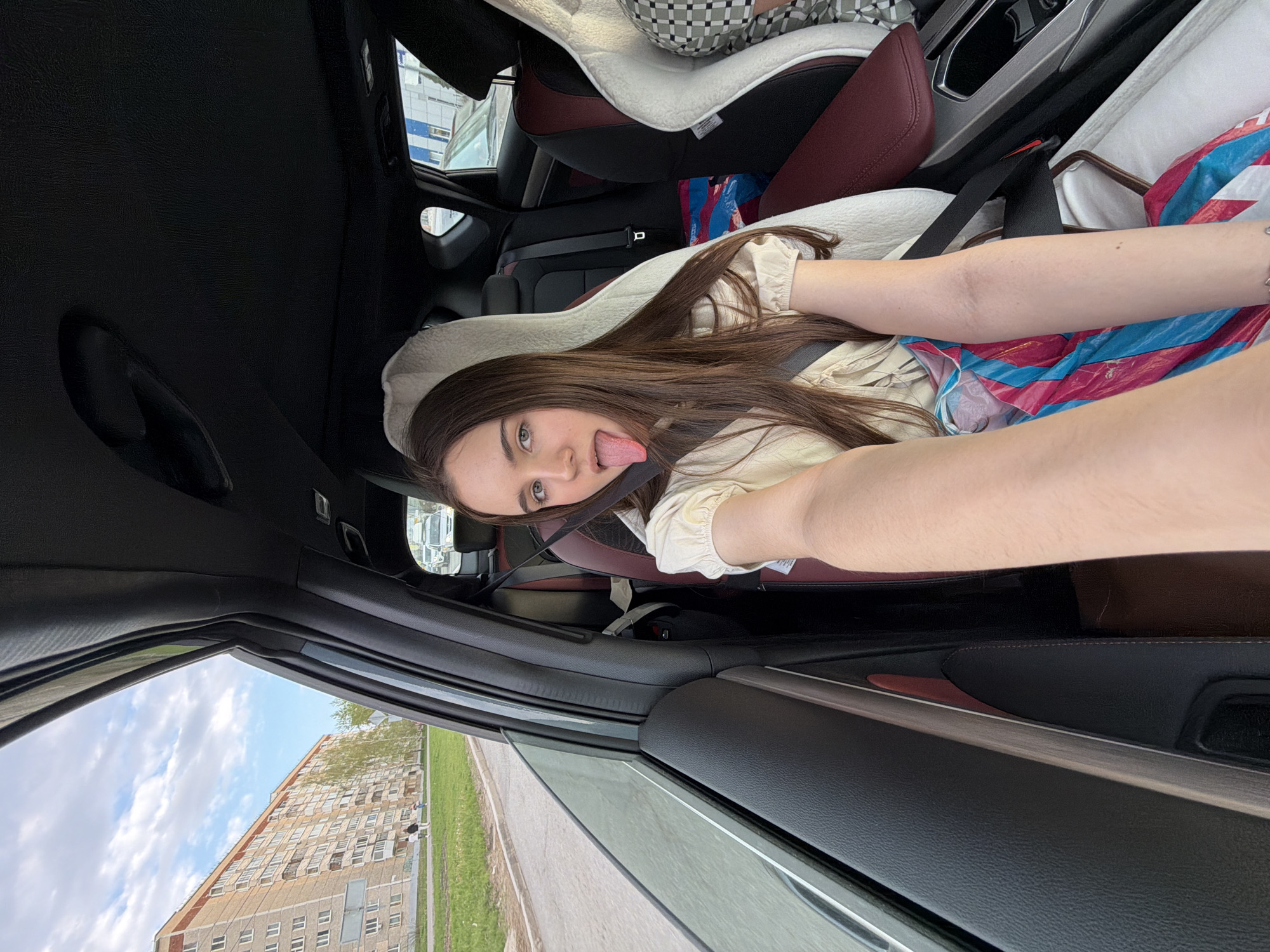 A woman is taking a selfie in a car. The interior of the car features red and black leather seats and a child safety seat. The woman is wearing a seatbelt and sticking her tongue out. Outside the car window, a multi-story residential building is visible in Izhevsk, Russia, with a grassy area and sidewalk nearby. The sky is partially cloudy.