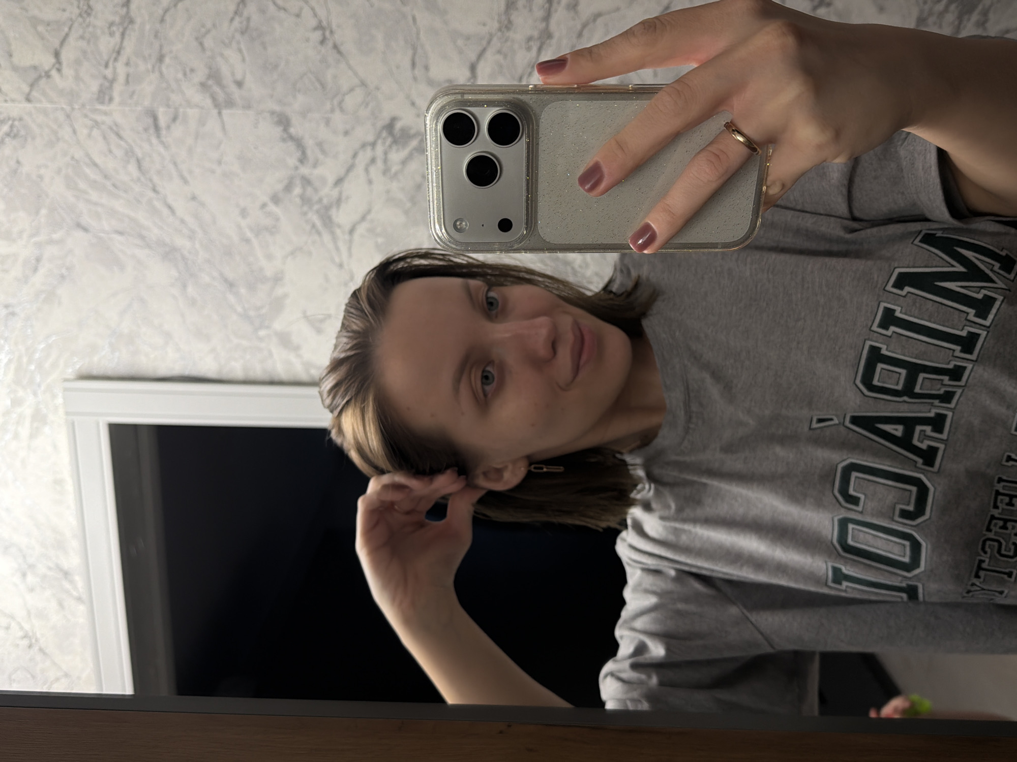 A young woman in Reshetnikovo, Russia, takes a mirror selfie indoors. She holds a smartphone with a clear, glitter-infused case in her right hand, showcasing its three prominent camera lenses. Her nails are painted a muted plum color, and a simple gold-toned ring adorns her right ring finger. She has shoulder-length brown hair, light-colored eyes, and a calm expression with a faint smile, looking towards the phone's camera.
Her left hand is raised, gently touching her left ear, revealing a small, gold-colored, chain-link earring. She is wearing a casual grey t-shirt featuring dark green, collegiate-style lettering. Although reflected and inverted, the text on her shirt appears to spell out "MIRACUL" or a similar word, followed by "COLLEGE" or "UNIVERSITY" on the right.
The background features a light-colored wall with a subtle grey marble pattern. To her left, a large, dark rectangular surface, possibly a television screen or a window, is framed by a white border. The bottom edge of the mirror she is using is a dark wooden surface. The lighting is soft and even, indicating an indoor setting with ambient light.