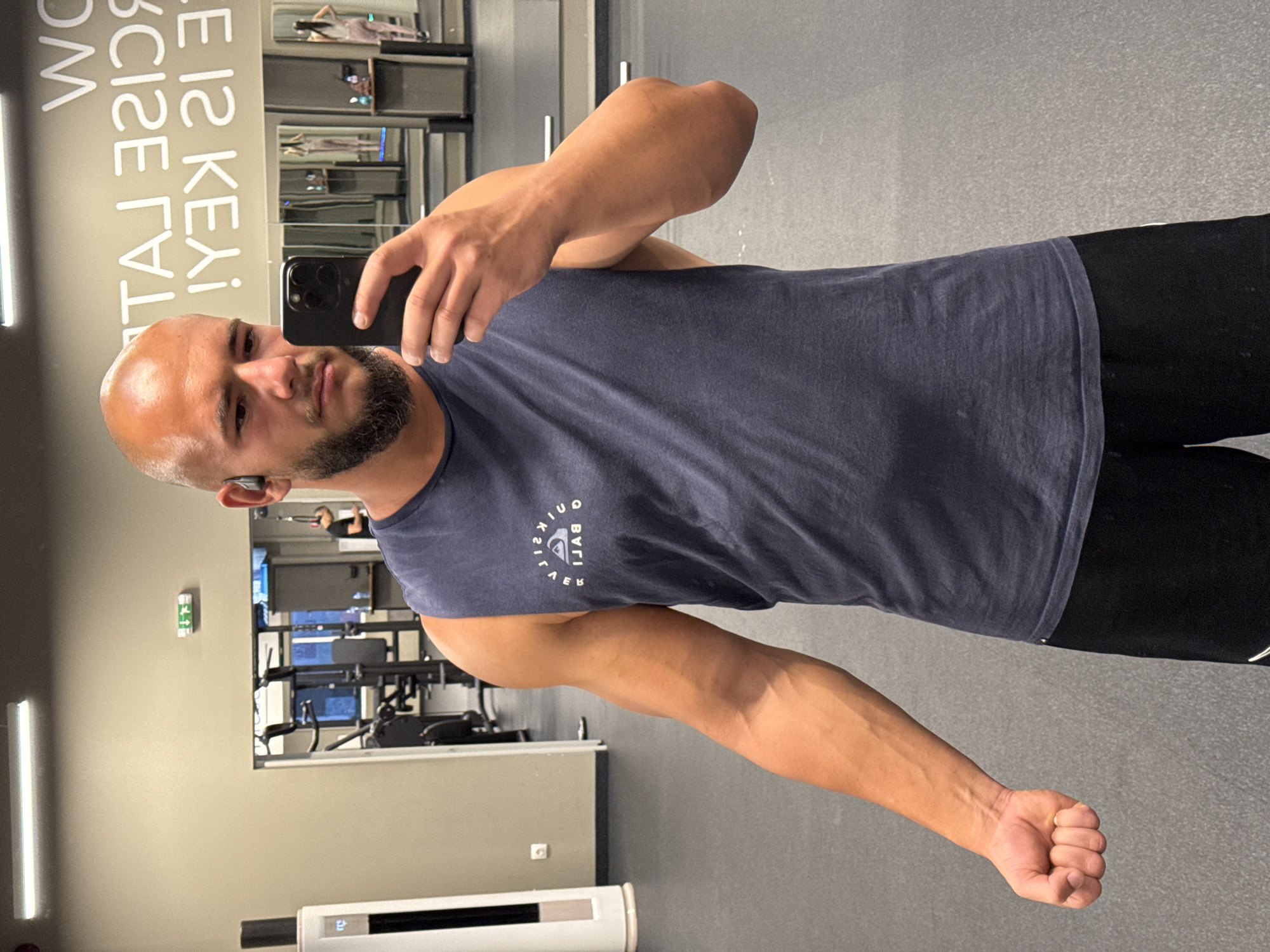 A bald, bearded man in a dark blue sleeveless shirt and black shorts is taking a selfie in a gym. He is holding a black smartphone in his right hand and wearing black wireless earbuds. In the background, there is gym equipment, mirrors, and a neon sign on the wall that reads "WORK IS KEY IS LATER". The floor is dark gray. The gym is located in Sofia, Bulgaria.
