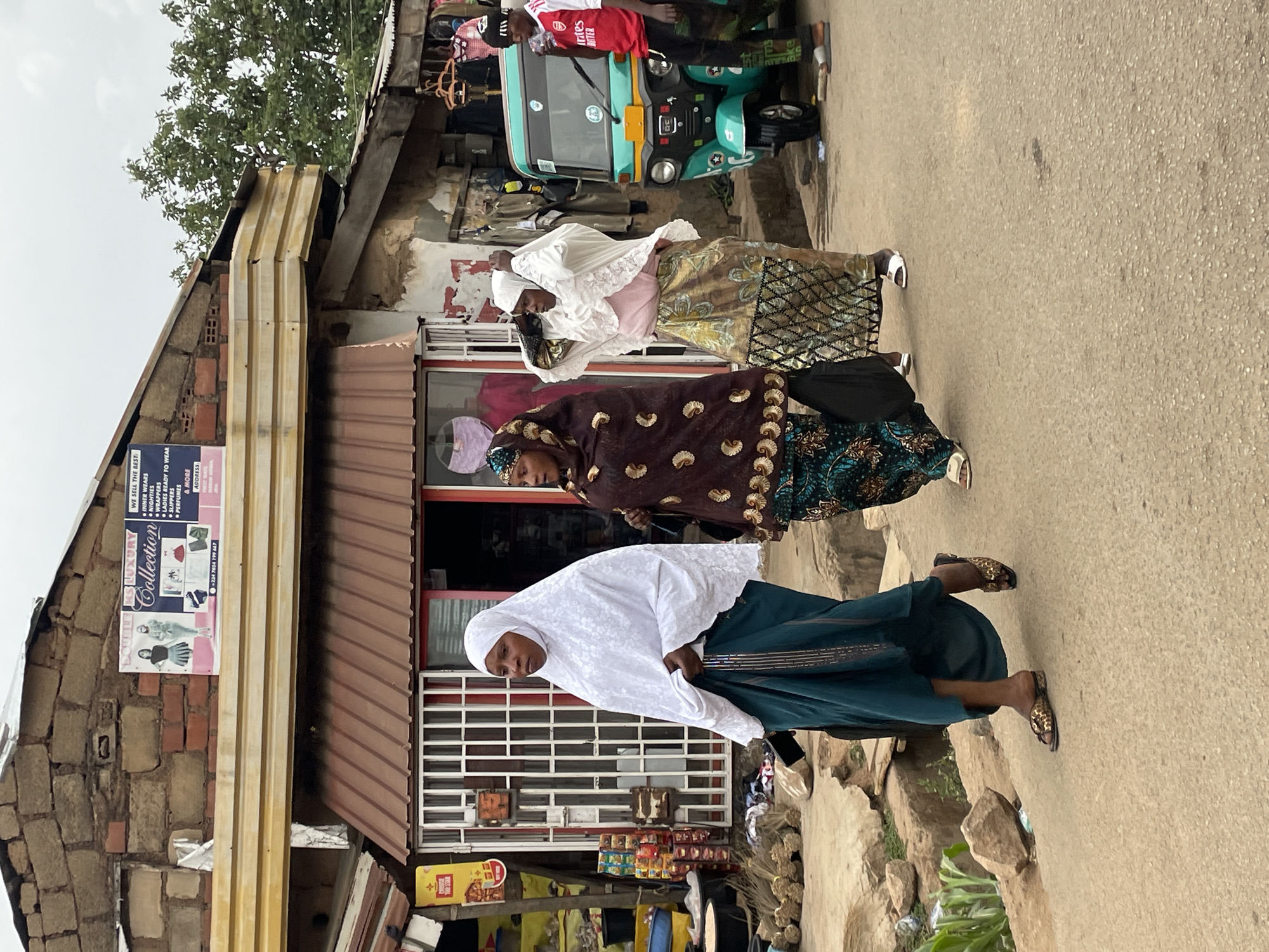 This daytime image captures a street scene in Jos, Nigeria, under an overcast sky. The street, made of worn asphalt, is populated by several pedestrians and lined by buildings featuring a mix of brick, concrete, and corrugated metal roofing.
In the foreground, a woman in a white hijab and a dark teal skirt with patterned sandals looks towards the viewer, standing in front of a shop with a security grille. Behind her, another woman walks away from the camera, dressed in a dark brown patterned top and green patterned skirt, carrying a black shoulder bag. Further down the street, a third woman also walks away, wearing a white, lace-trimmed top and a gold-toned patterned skirt.
In the background, a person wearing a red t-shirt (displaying "BETTER" and "intes" from an Arsenal FC jersey), dark pants, and a cap stands next to a bright green motorized tricycle, a common mode of transport known as a keke napep. The tricycle also has a red jerrycan or container visible at its front.
The buildings on the left side of the street house various businesses. A prominent sign for "DOUBLE R Luxury Collection" is affixed to a brick wall, advertising "INNER WEARS," "WRAPPERS," "LADIES READY TO WEAR," "SLIPPERS," "PERFUMES," and "& MORE," along with a phone number: "+234 7034 199 647." Other smaller signs and goods, such as brooms and packaged items, are visible outside the shops. The scene depicts typical daily life in a bustling local area.