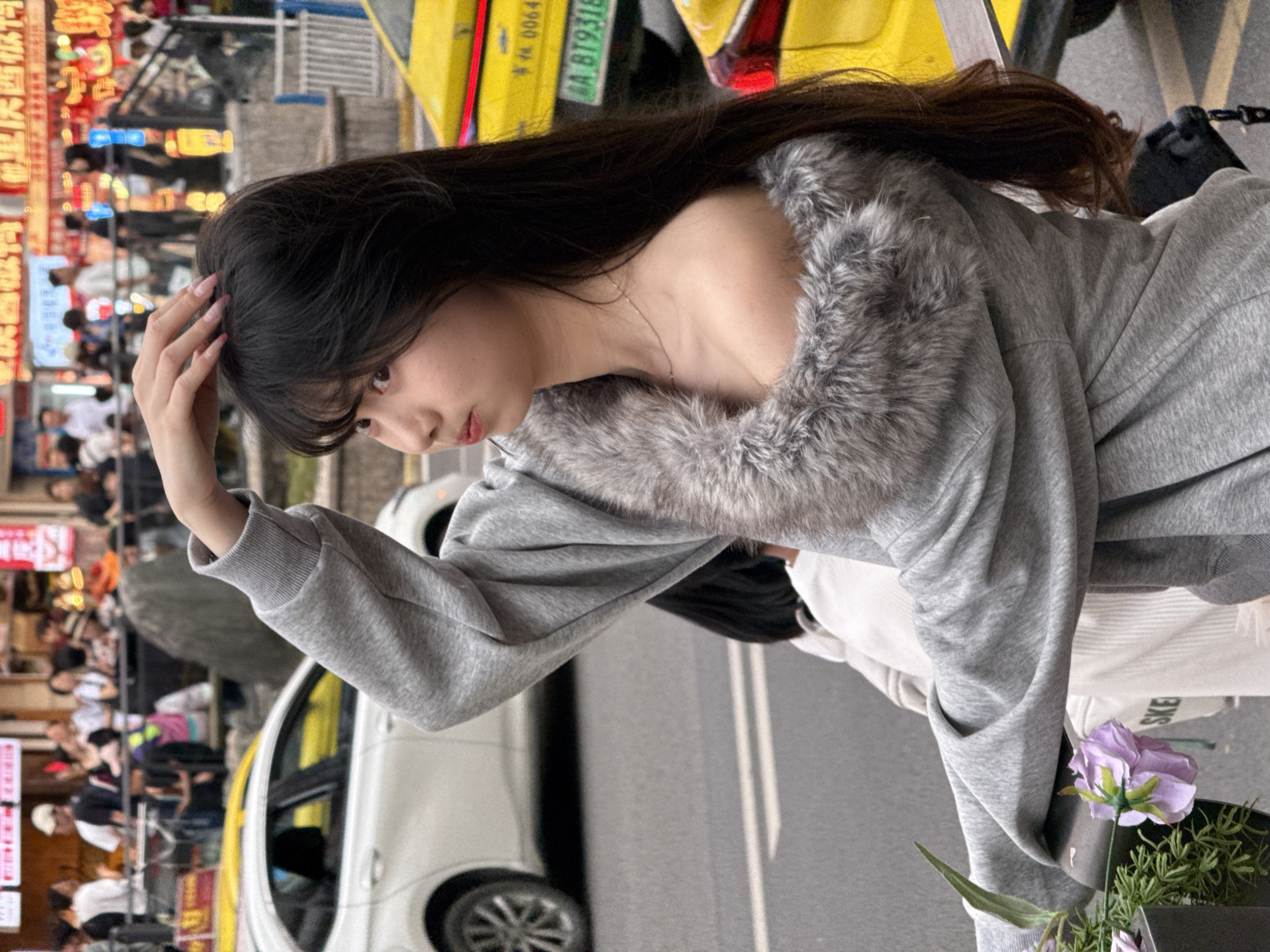 A young woman stands on a city street in Liangjiang, China, holding her hair with her right hand. She is wearing a grey sweatshirt with a faux fur collar. In the background is a white sedan, a yellow taxi, street signs in Chinese, and pedestrians. A small pot with purple flowers and greenery is in the lower right.