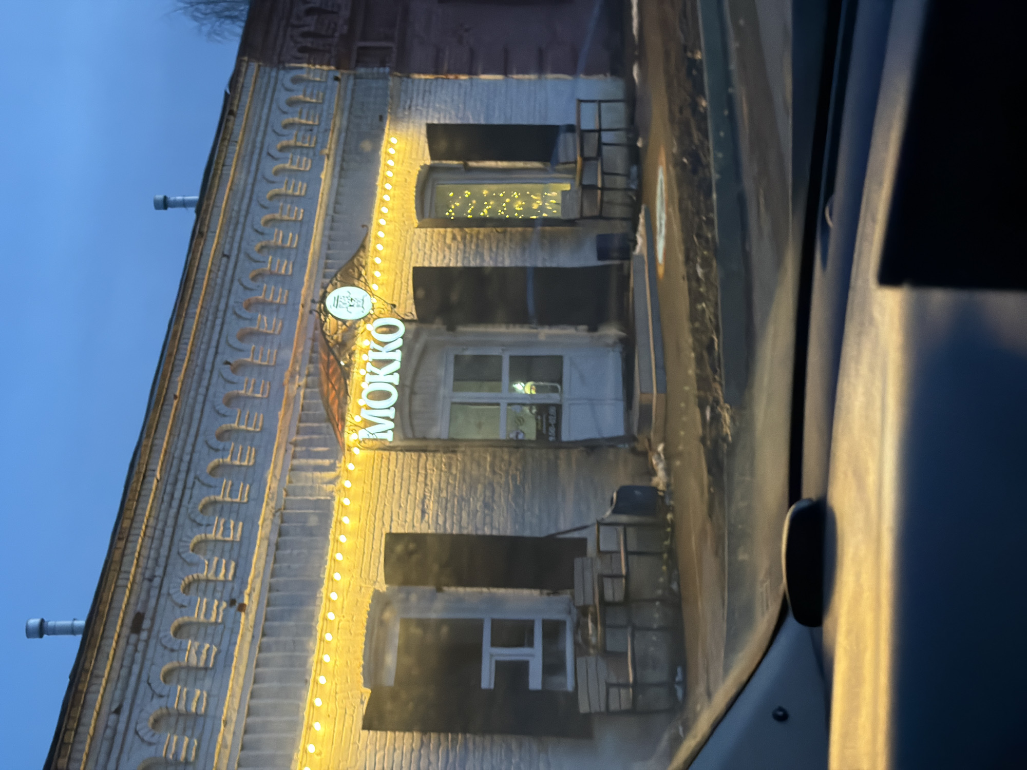 Captured through a car window, the facade of a brick building is illuminated by string lights framing its vertical length. Above the entrance is the word "MOKKO" displayed in vertically aligned, illuminated letters. The building, likely a commercial establishment, features shuttered windows. Located in Sarapul, Russia, the setting is likely urban, evidenced by the architectural design and commercial signage. The car's interior and window frame parts of the composition. The time of day appears to be evening, indicated by the twilight sky.