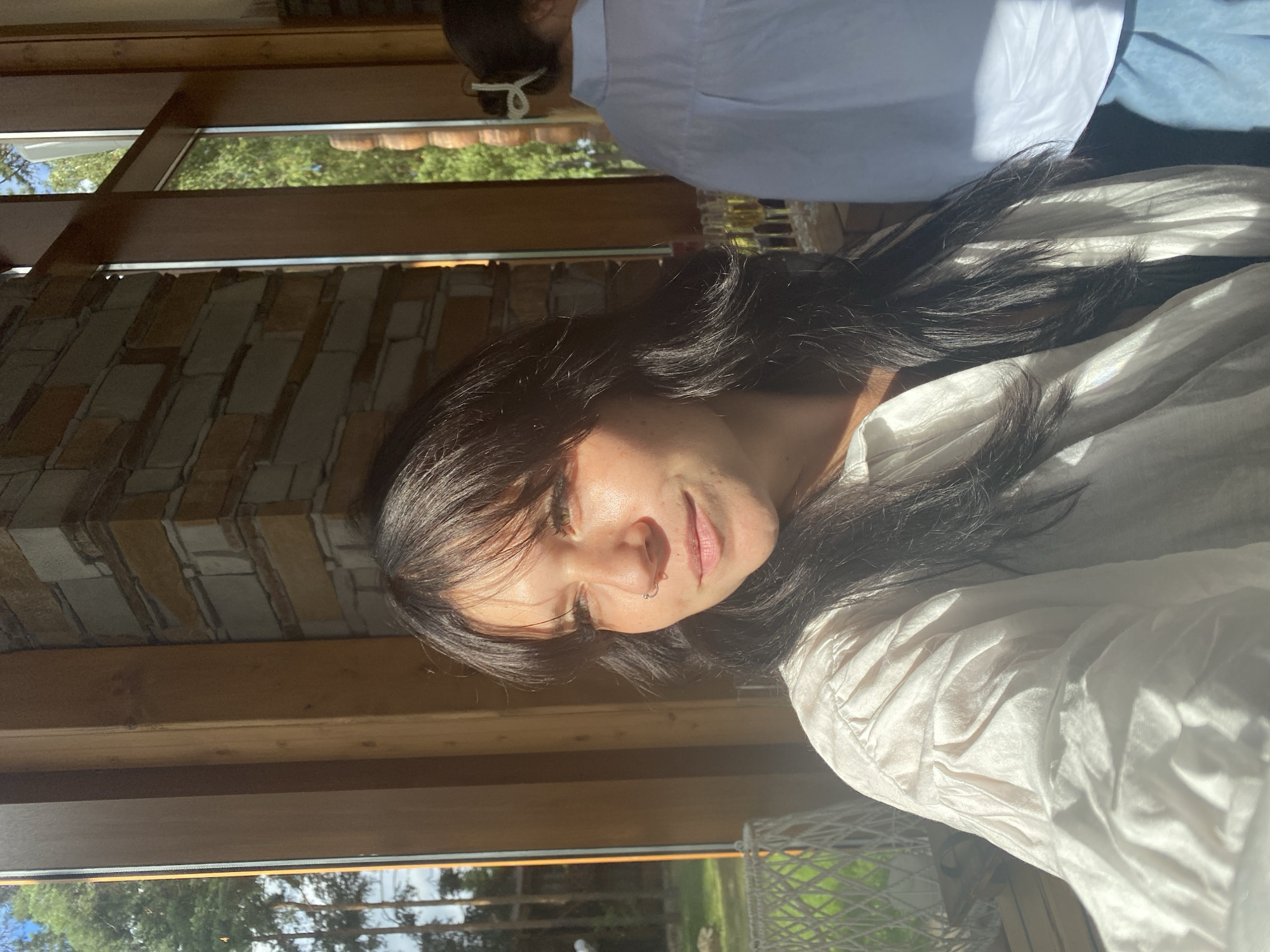 A woman with long, dark hair is shown in a close-up shot, looking to her left. She is wearing a light-colored, loose-fitting shirt. The background features large windows with wooden frames, a brick wall, and glimpses of trees and greenery, suggesting an indoor location with an outdoor view. The lighting indicates it is daytime, possibly late afternoon given the warm sunlight. The overall setting appears to be a contemporary building, possibly a cafe or a residential space, with a naturalistic architectural style, consistent with locations in Krasnoflotskoe, Russia. Another person wearing a light blue shirt is visible in the background, out of focus.