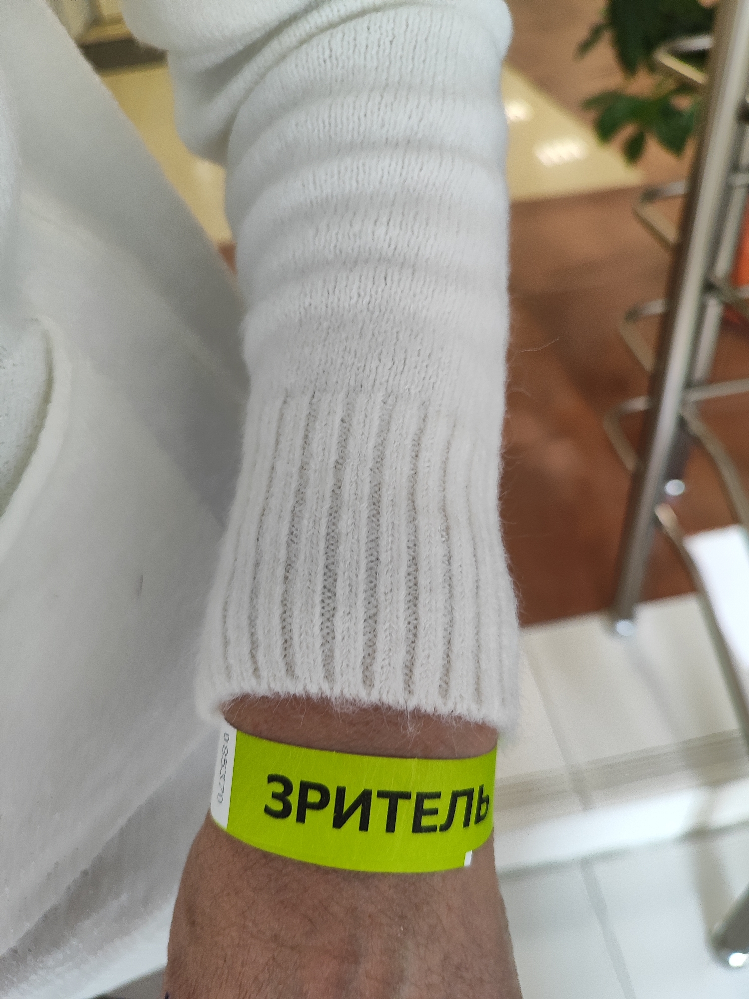Here's a detailed description of the image:
A person's left arm is prominently featured, wearing a white, thick-knit sweater with a ribbed cuff that ends just above the wrist. Around the wrist, there is a bright yellow wristband with the Russian word "ЗРИТЕЛЬ" written on it in black capital letters. The skin on the wrist is visible below the band.
The background shows a blurred interior setting, possibly indoors due to the neutral colored walls, suggesting the location might be a public event in the city of Fryazino, Russia. A plant and parts of metal-framed chairs are visible in the out-of-focus background.