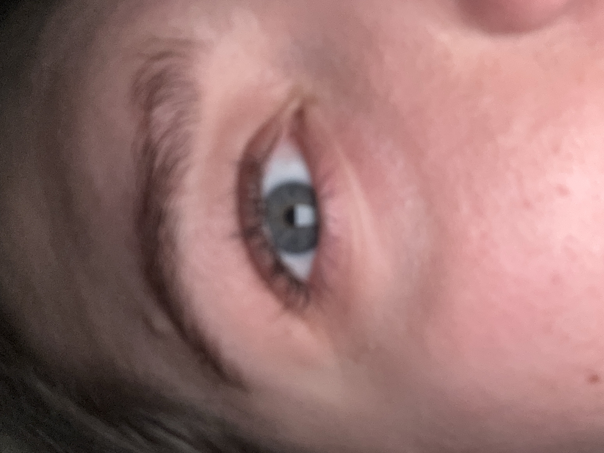 The image is a close-up of a human eye with blue-gray irises. The skin tone is fair with some reddish undertones, and there are visible pores and slight texture. The eyebrow above the eye is dark and appears to be naturally shaped. The hair on the left side of the image is dark brown and slightly blurred, suggesting it is out of focus. The background is also blurred, with variations in pinkish and brownish tones. There is no discernible location in Saratov, Russia, or any specific environmental details provided by the image itself. The image focuses solely on the eye and surrounding facial features.