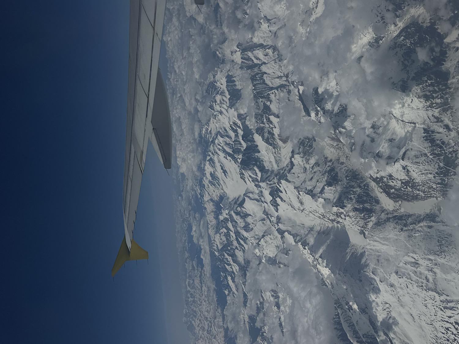 This aerial photograph captures a breathtaking view from an airplane, showcasing a partial aircraft wing against a backdrop of vast, snow-capped mountains and a clear blue sky. The scene is observed from a high altitude, characteristic of an airborne journey.
In the foreground, the left wing of a commercial airplane extends from the mid-left of the frame, angling upwards towards the top-left corner. The wing is predominantly light grey, with visible panel lines and a distinctive yellow fin or wingtip device at its outermost point.
The main subject in the background is an expansive and rugged range of snow-covered mountains, occupying the right two-thirds of the image. The peaks and valleys are heavily blanketed in white snow, with darker rock faces and shadows providing stark contrast. Scattered patches of white and grey clouds are visible among the mountain peaks, adding depth and texture to the scene. The sky above the mountains and to the left of the wing is a deep, clear blue, suggesting excellent visibility and bright daytime conditions. The horizon where the sky meets the distant land appears hazy, typical of views from high altitudes.
There are no people or visible text in the image. The time of day is clearly daytime, given the bright illumination. The extensive and deeply snow-covered mountains suggest a flight over a major mountainous region.