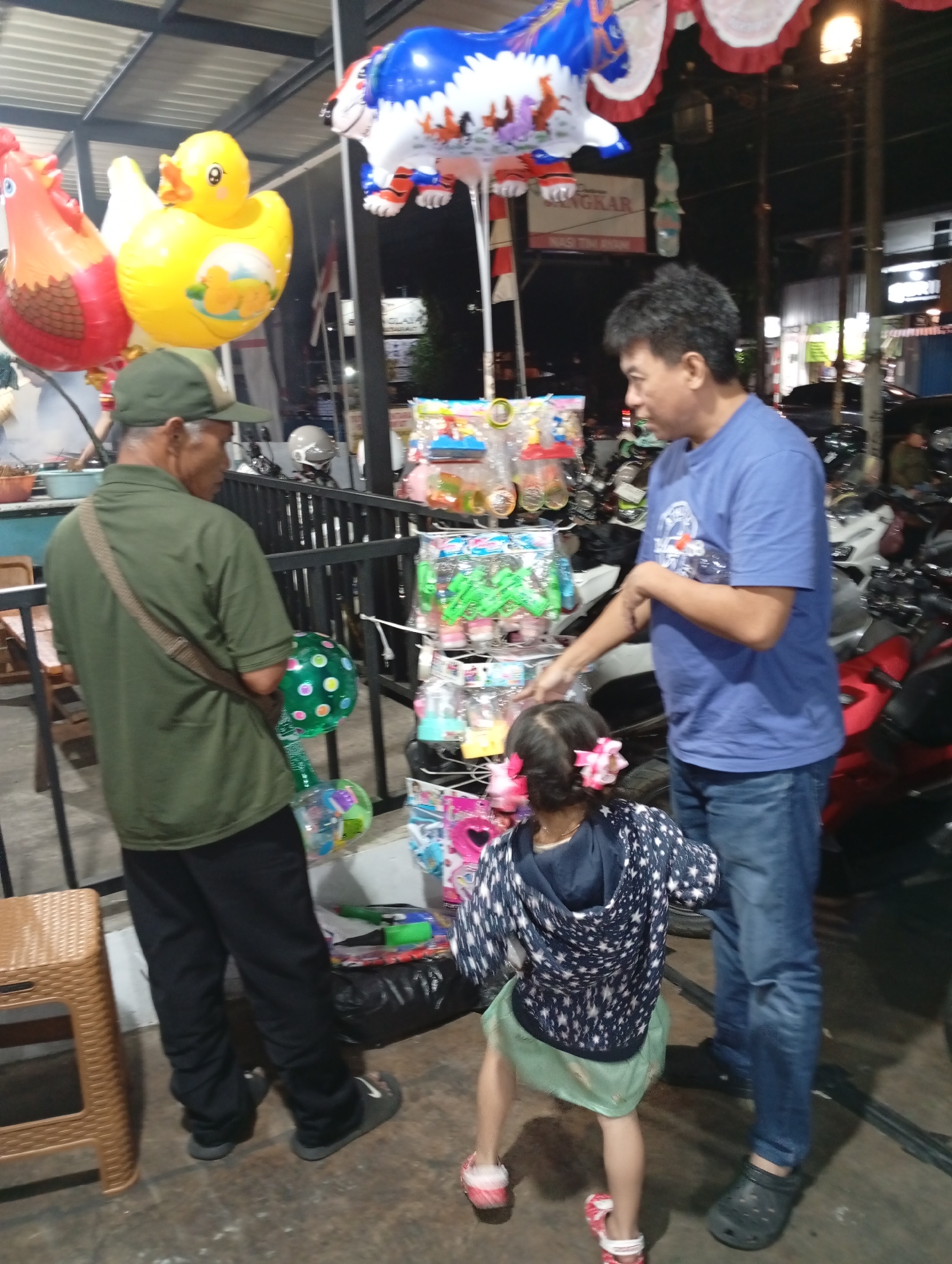 At night, in a street-side stall area in Pacet, Indonesia, a man in a blue t-shirt and jeans stands interacting with a young girl in a patterned hoodie and a green skirt. The man is pointing at a display of colorful balloons and toys. To the left, an older man in a green shirt and a baseball cap is looking away from the camera, with a green balloon in his hand. Behind them, various motorcycles and scooters are parked. Several large, inflated balloons, including a yellow duck, a red rooster, and a blue and white tiger, are hanging above the stall. The environment appears to be an outdoor market or roadside area.