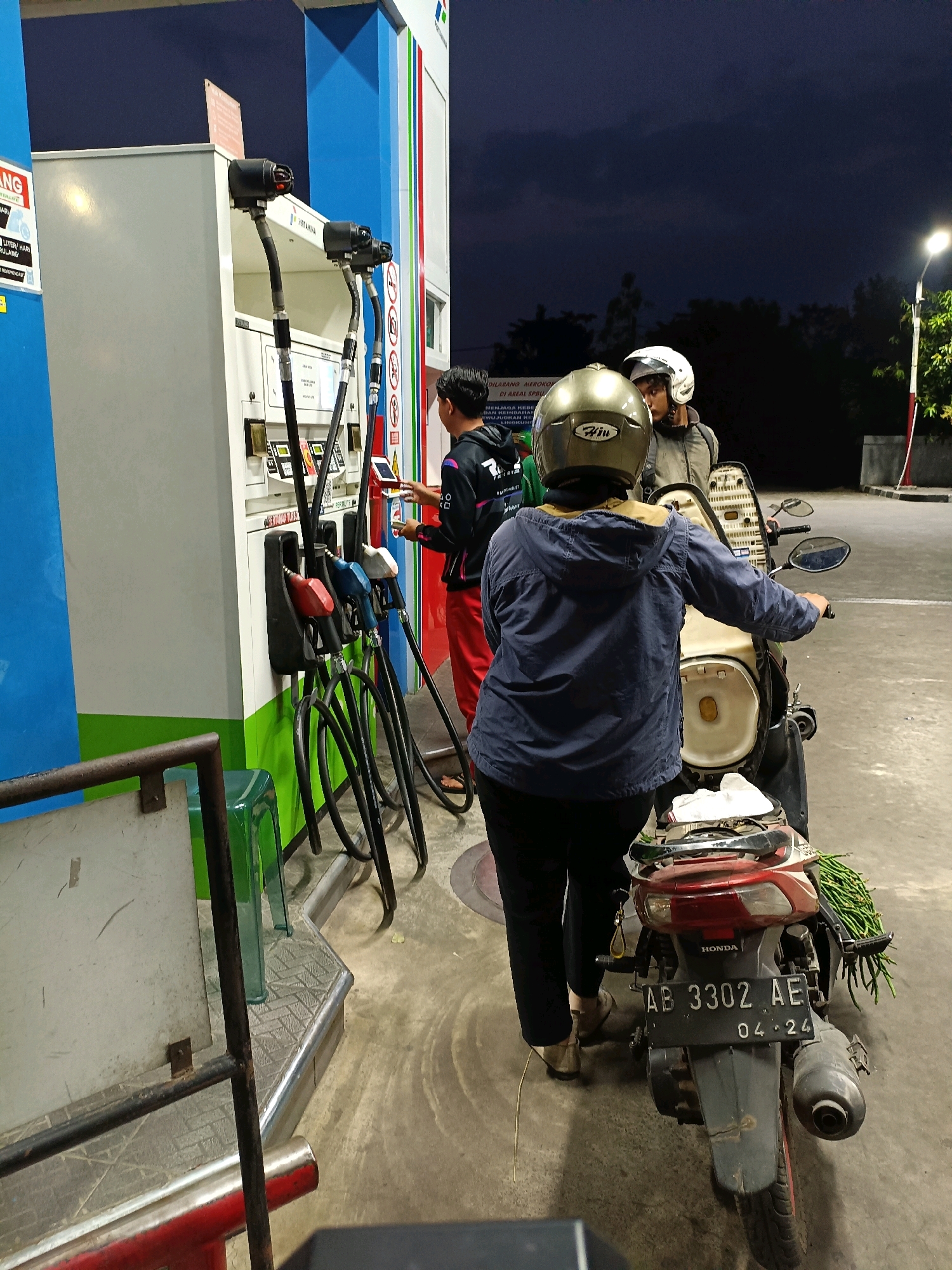 At a Pertamina gas station in Maguwoharjo, Indonesia, two individuals are at a fuel pump at dusk. One person, wearing a blue jacket and a gold helmet, is seated on a motorcycle. The other person, wearing red pants and a black hoodie with white accents, is standing and interacting with the fuel pump's touchscreen. A third person, wearing a white helmet and a light-colored jacket, is also on the motorcycle, facing away from the camera. The motorcycle's license plate reads "AB 3302 AE" with "04-24" below it. The gas pump has several hoses with different colored nozzles. The environment is a paved area with a dark sky overhead, suggesting it is nighttime or late evening.