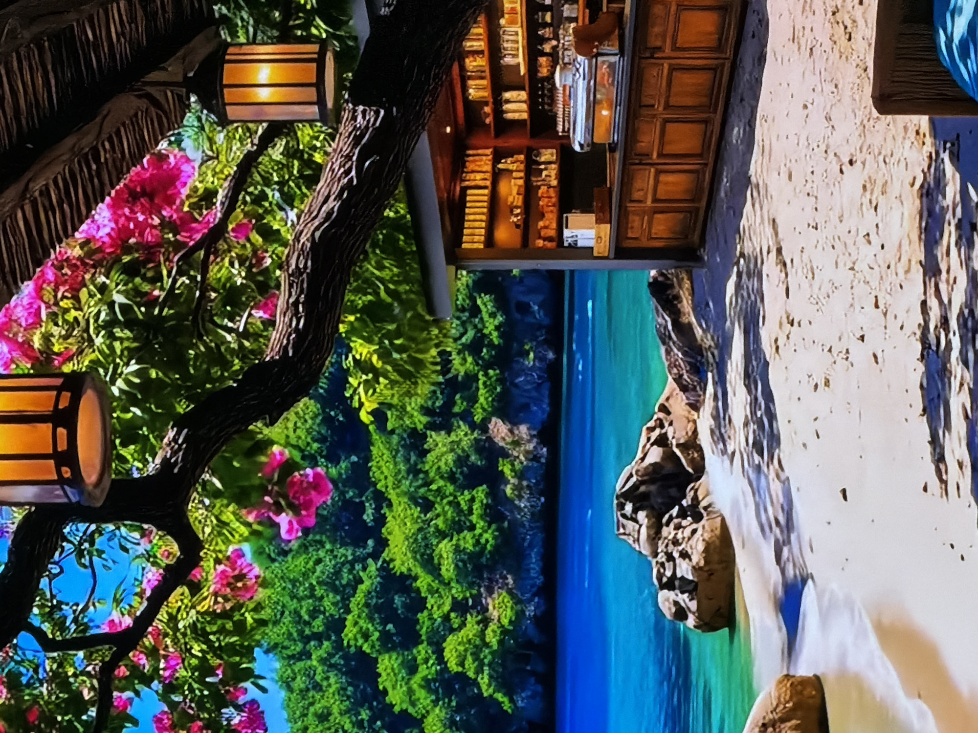 The image displays a vibrant, picturesque scene resembling a tropical beach setting, possibly a large-scale digital display or a mural, captured with a slight grainy texture. The orientation is currently rotated 90 degrees clockwise, so for description, we'll consider the beach on the right, the water in the middle, and lush greenery with structures on the left.
On the left side, a dark, gnarled tree trunk and branches extend into the frame, adorned with bright pink, bougainvillea-like flowering plants and dense green foliage. Attached to the tree structure are two glowing wooden lanterns, providing a warm, inviting light with horizontal slats. Partially visible at the very top left is a section of a thatched roof. Further to the left, nestled among the foliage, is a well-stocked wooden bar or counter. Its shelves are filled with bottles, glasses, and various items, indicating a leisure establishment.
The midground features a band of dark green, dense tropical foliage that transitions into calm, clear turquoise and blue ocean waters. Rocky formations, some covered with light growth, line the edge where the water meets the land.
On the right side, a wide expanse of light-colored sandy beach is visible, textured with ripples and shadows. A blue and white patterned object, possibly a cushion or towel, is partially seen at the far top right of the beach.
No people are visible in the scene. The warm glow of the lanterns suggests either evening or nighttime, or a mood-lit indoor environment. The tranquil water and lush vegetation evoke a sense of relaxation and a vacation atmosphere. Despite the provided context of Sofiivska Borshchahivka, Ukraine, the depicted scene strongly portrays a tropical coastal environment. No visible text is present in the image.