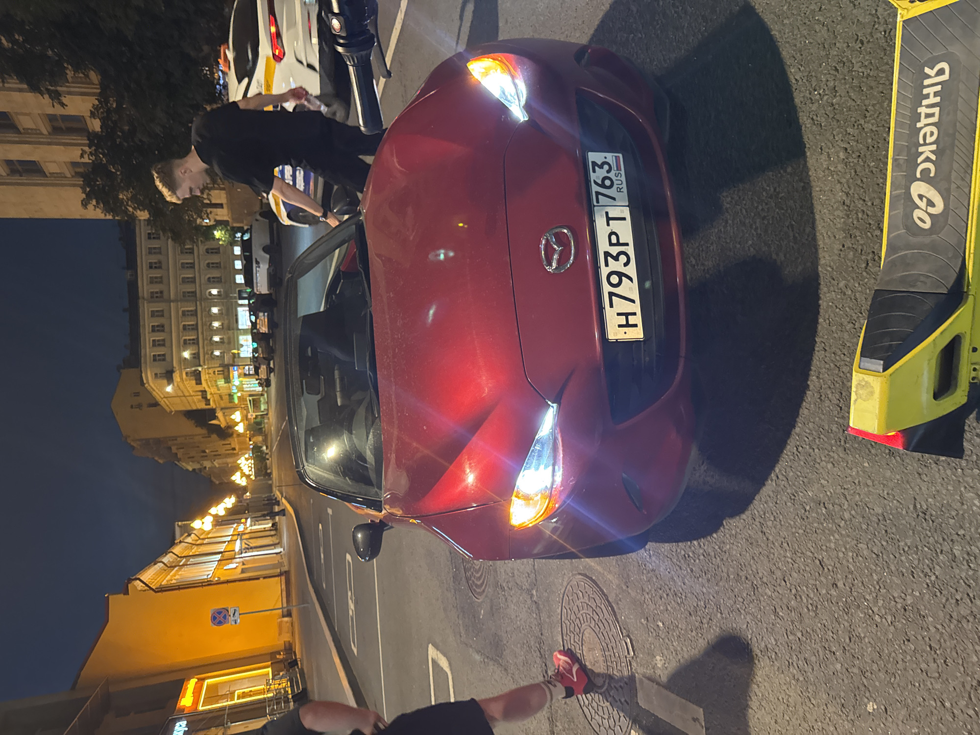 The image depicts a street scene at night in Moscow, Russia. In the foreground, a red Mazda convertible is parked, its headlights illuminated. A man in a black t-shirt and dark pants stands near the driver's side of the car. To the right, a white vehicle, possibly a police car given the orange light on top, is partially visible. In the lower portion of the frame, a yellow electric scooter with the "Yandex Go" logo is lying on the asphalt. The street is lined with buildings, and streetlights cast a warm glow. The overall impression is of a street in a city at night, with urban elements and vehicles present.