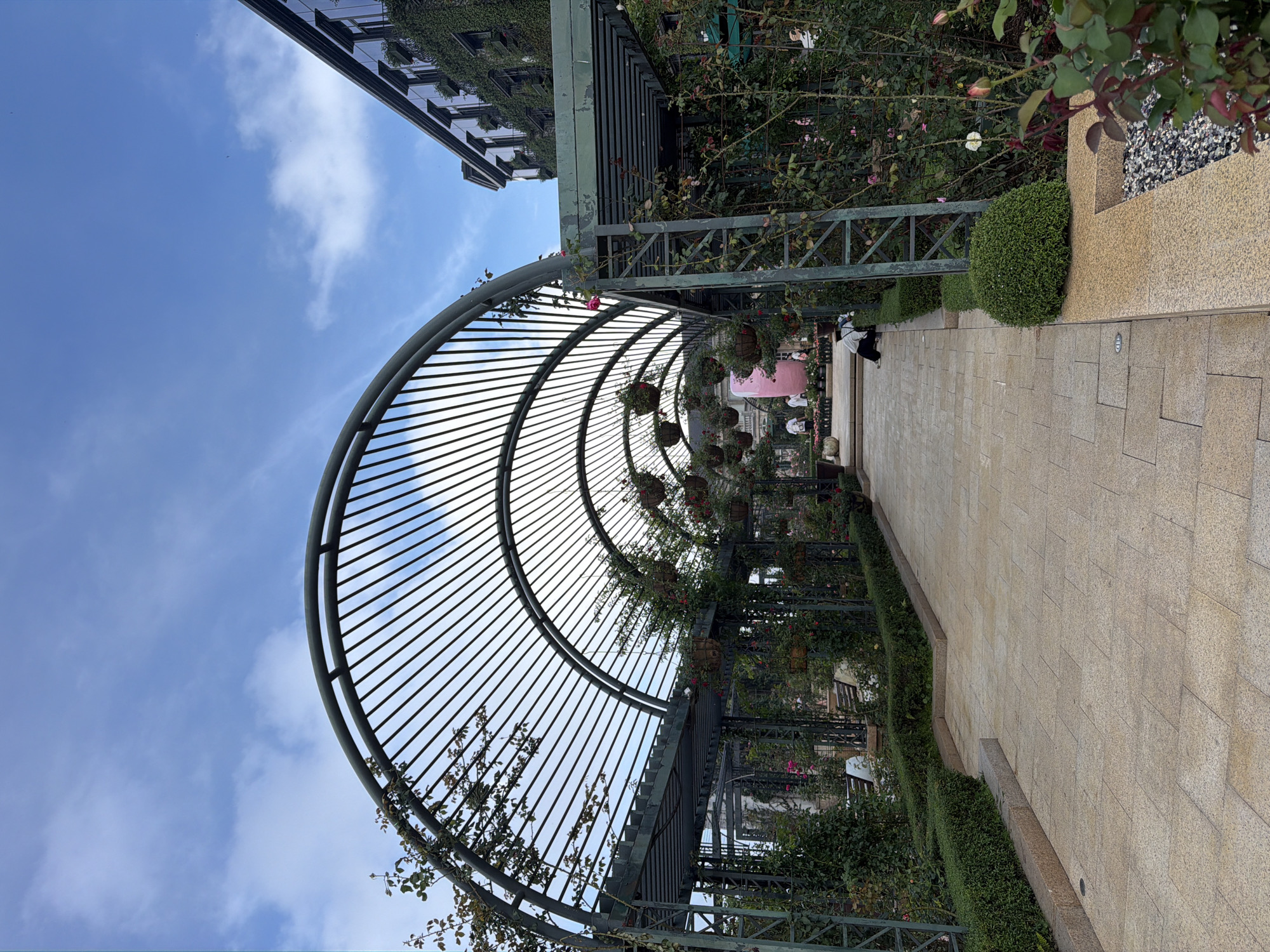 The image depicts a stone-paved pathway beneath a metal-framed trellis in what appears to be a landscaped park. The trellis is adorned with hanging flower baskets and climbing vegetation. The path is lined with manicured hedges and rose bushes. In the distance, there are a few individuals walking along the path. The architecture suggests a modern urban environment with a building visible partially in the background. Based on the architectural style and the presence of lush vegetation, it is plausible that this scene is located in Đà Nẵng, Vietnam.