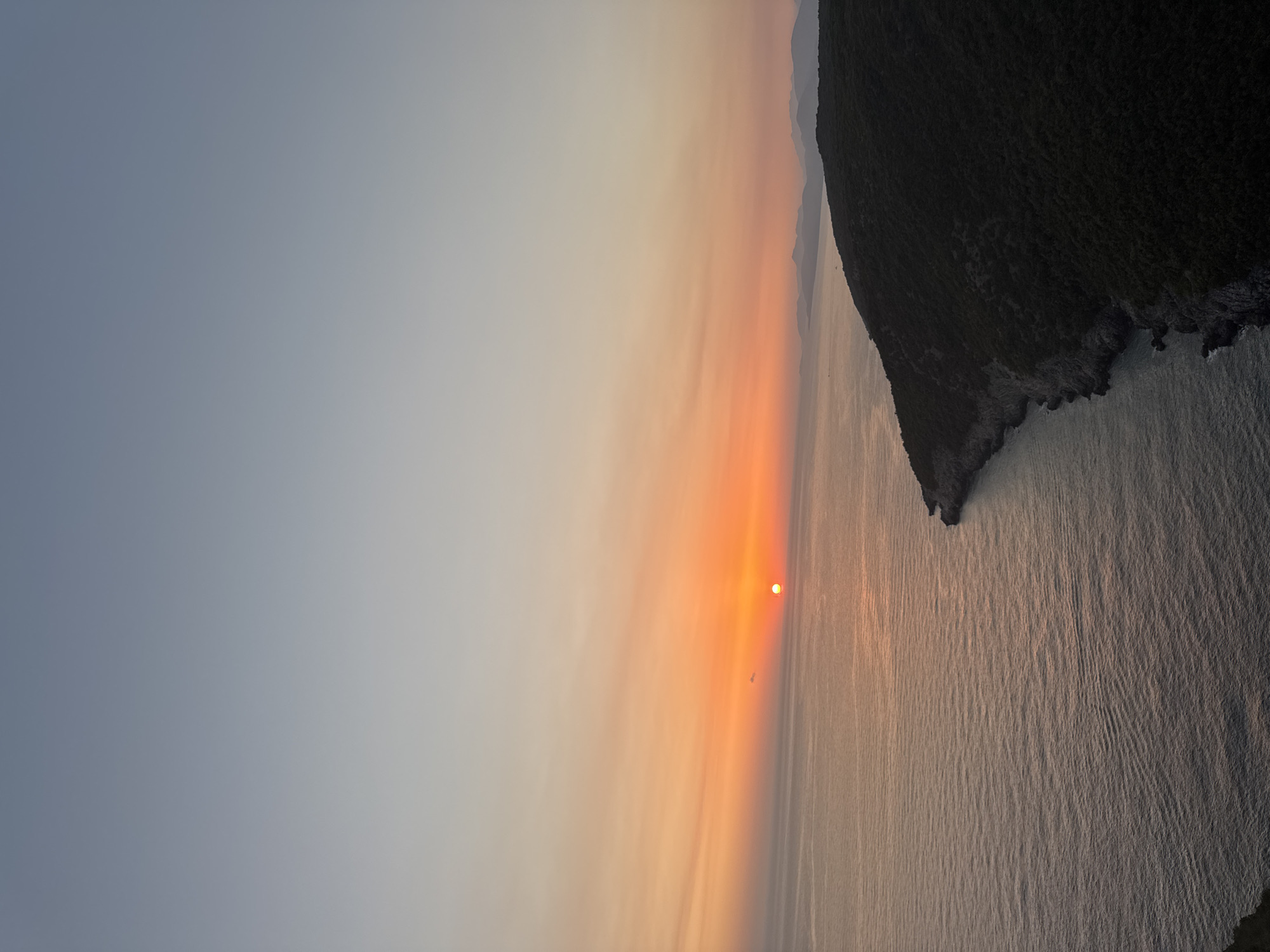 The image depicts a sunset over the Adriatic Sea, viewed from a high vantage point on the coast. The sun, a small, bright orange orb, is just touching the horizon, casting a warm glow that transitions into shades of orange and pink in the sky above. The sea is a deep blue, with gentle ripples reflecting the colors of the sunset. A dark, tree-covered headland juts out into the water on the right side of the frame, its rocky shoreline meeting the sea. In the distance, a hazy, mountainous coastline is visible. The sky is a clear gradient of pale blue at the top, transitioning to softer hues of orange and yellow near the horizon. There are no people or man-made structures clearly visible in the scene, suggesting a natural and remote landscape. The lighting is soft and diffused, characteristic of the golden hour. The location is the city of Zagora, Montenegro.