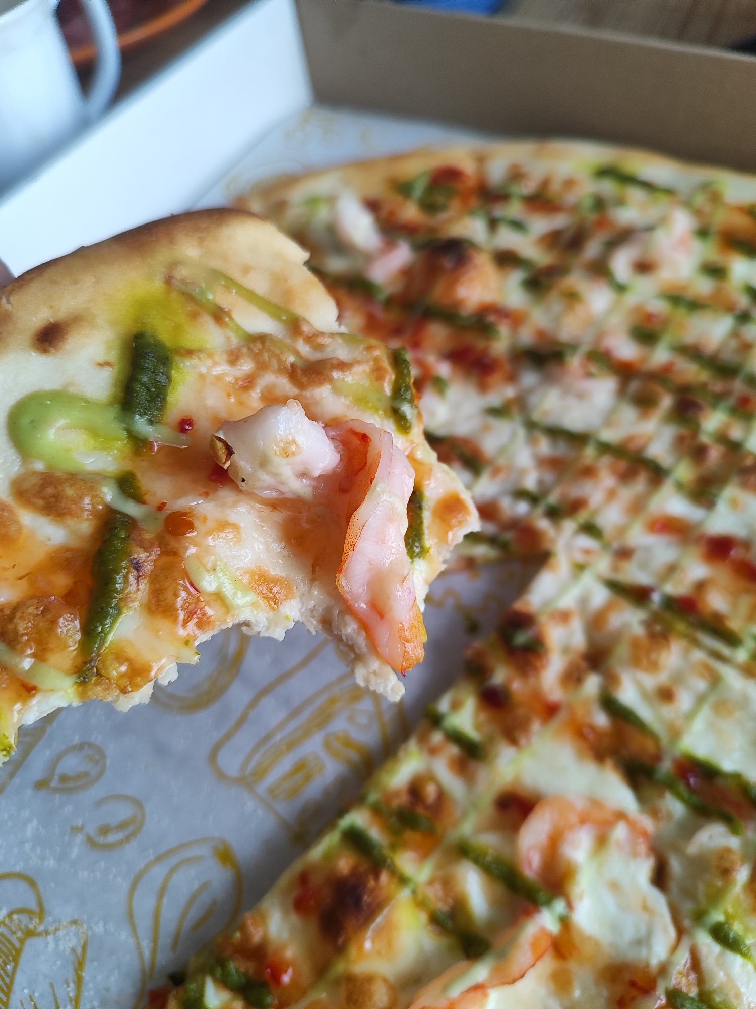 The image displays a close-up, high-angle view of a shrimp pizza in a white cardboard box, captured in Marusino, Russia. A single pizza slice is elevated and held by an unseen hand, revealing a bite mark near its center. A cooked, pink shrimp is prominently featured on the bitten edge of the elevated slice. The slice's toppings consist of melted white cheese, streaks of orange-red oil or sauce, and distinct lines and dots of vibrant green pesto. Small red chili flakes are scattered over the cheese. The crust is golden-brown.
The larger portion of the pizza, remaining in the white cardboard box, is slightly out of focus. It shows a similar arrangement of toppings: scattered pink shrimp, melted white cheese, orange-red sauce, and green pesto drizzled across its surface. The pizza is cut into square or rectangular segments. The interior of the white cardboard box is visible, lined with white paper that exhibits subtle yellow line-art graphics.
In the blurred background, a segment of a white ceramic mug or cup handle is visible on the far left. A generalized brown surface, likely a table, occupies the upper right background area.