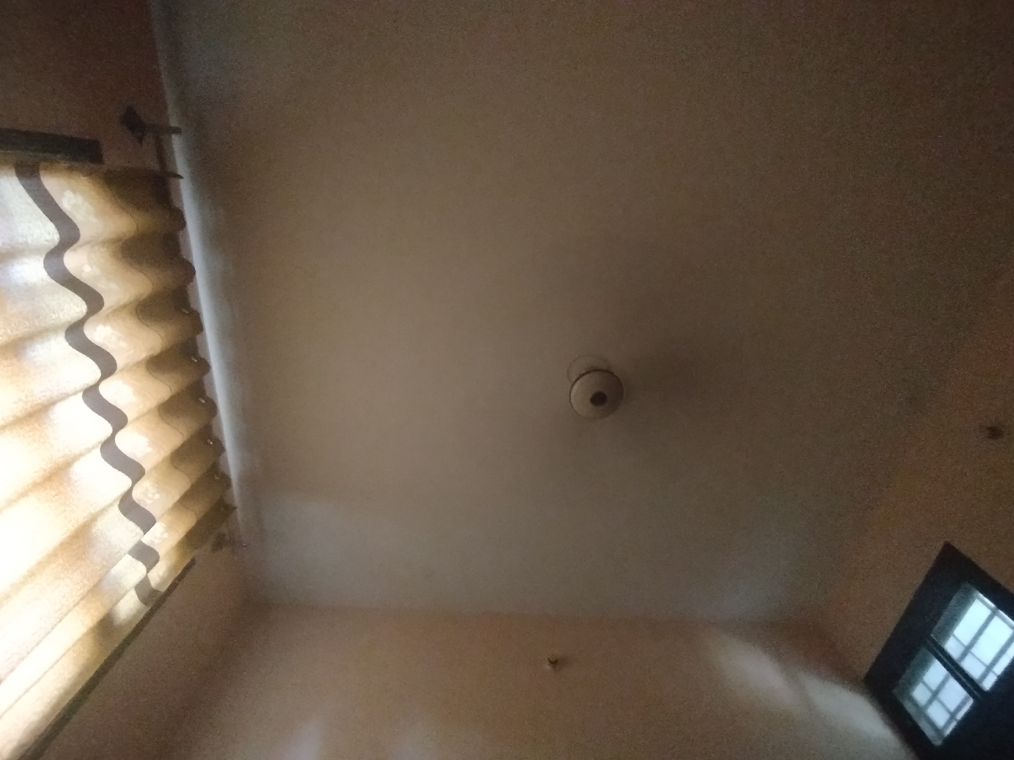 The image shows an interior view looking upwards towards a ceiling and a wall. On the left, there are pleated curtains with a wavy pattern, illuminated by natural light. In the center of the ceiling, there is a round, light-colored object, likely a ceiling light fixture or smoke detector. On the right side, a portion of a window with dark frames and multiple panes is visible. The walls and ceiling appear to be painted in neutral, light colors. The scene is domestic in nature and does not provide any specific information about the city of Lassan, Pakistan.