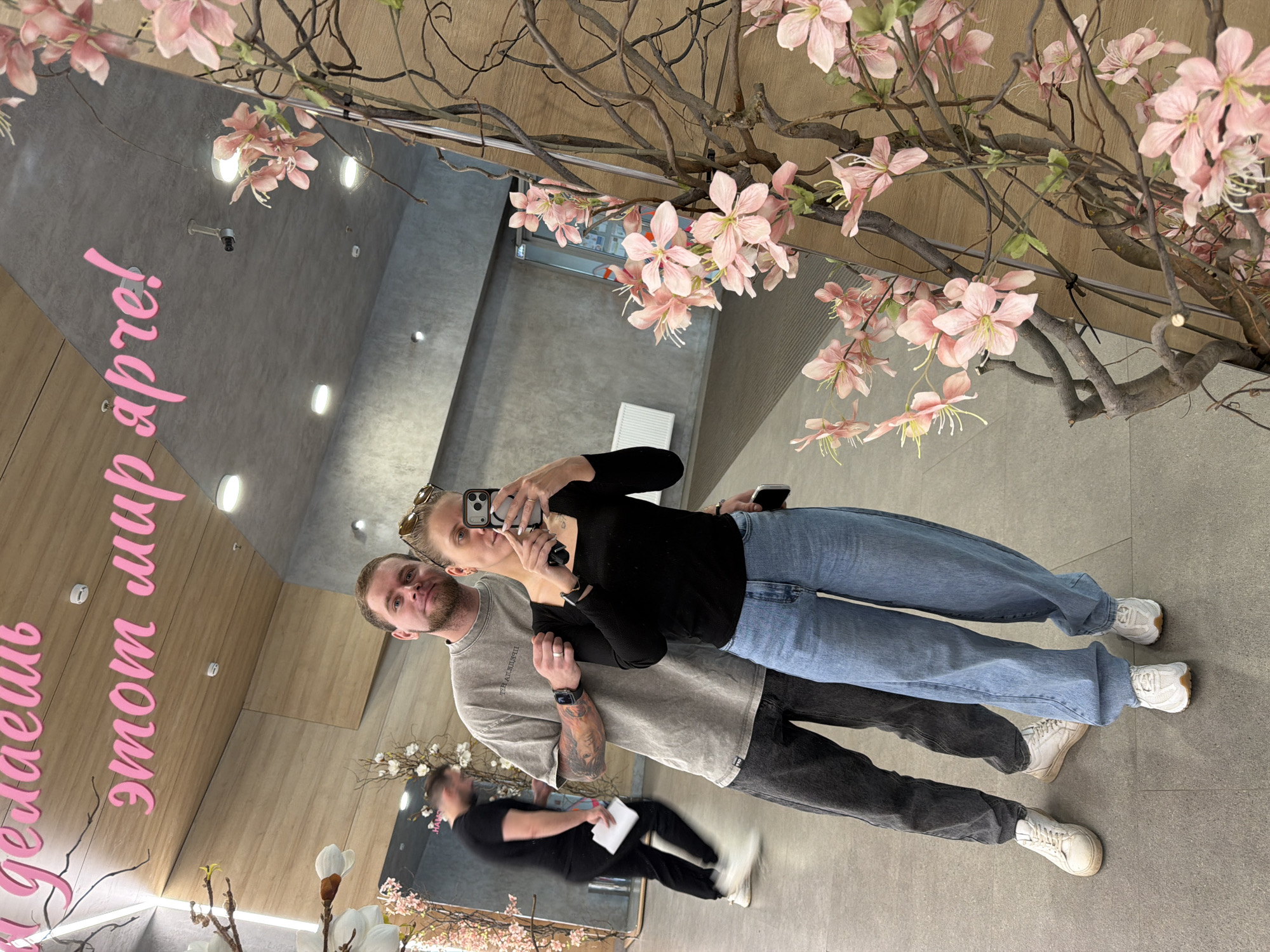 A couple is taking a selfie in a mirror, capturing a reflection of the interior. The location appears to be in Izhevsk, Russia, as indicated by the text on the mirror "делаешь этот мир апреле!". The woman is wearing a black long-sleeved shirt and blue jeans, while the man is wearing a gray T-shirt and dark gray jeans. The woman is holding a smartphone, presumably taking the picture. A person is seen in the reflection behind the couple. The interior features decorative elements including flowering branches and a gray wall.