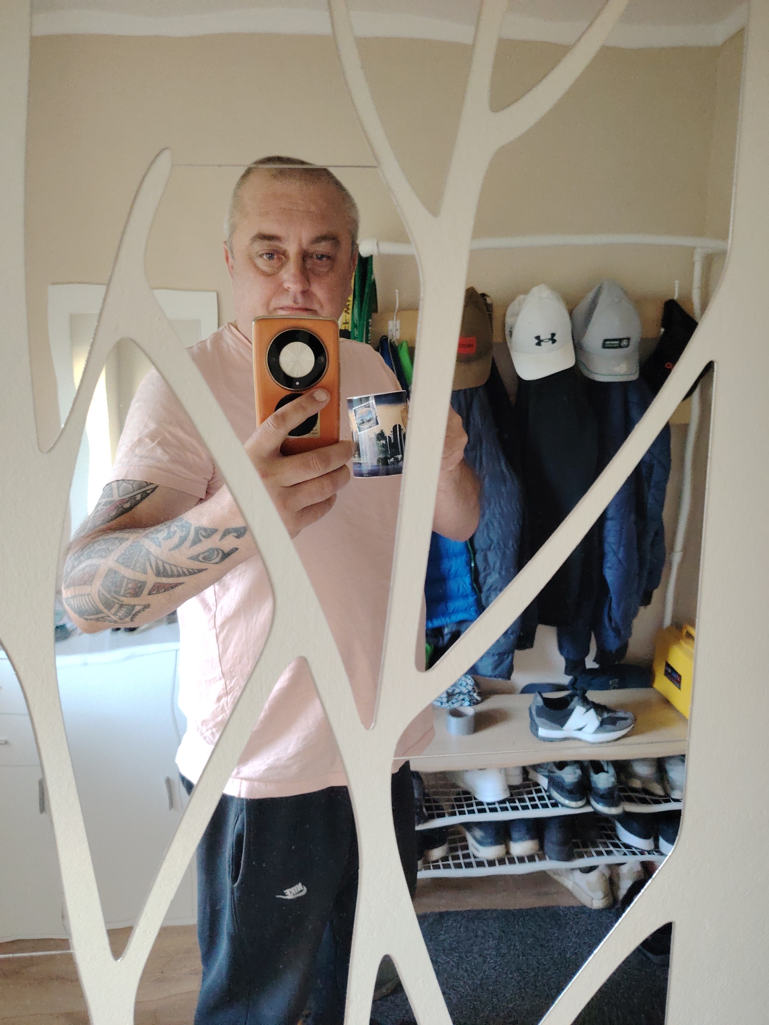 The image shows a man in his 40s taking a selfie in a mirror. He is wearing a light pink t-shirt and black sweatpants with a white Nike logo on the left leg. He has tattoos on his left arm and is holding an orange smartphone in his right hand. In the background, there are several coats and hats hanging on a rack, and a shoe rack with multiple pairs of shoes. The room appears to be an entryway or mudroom. There are no discernible features that place this image in Huntingdonshire, United Kingdom.