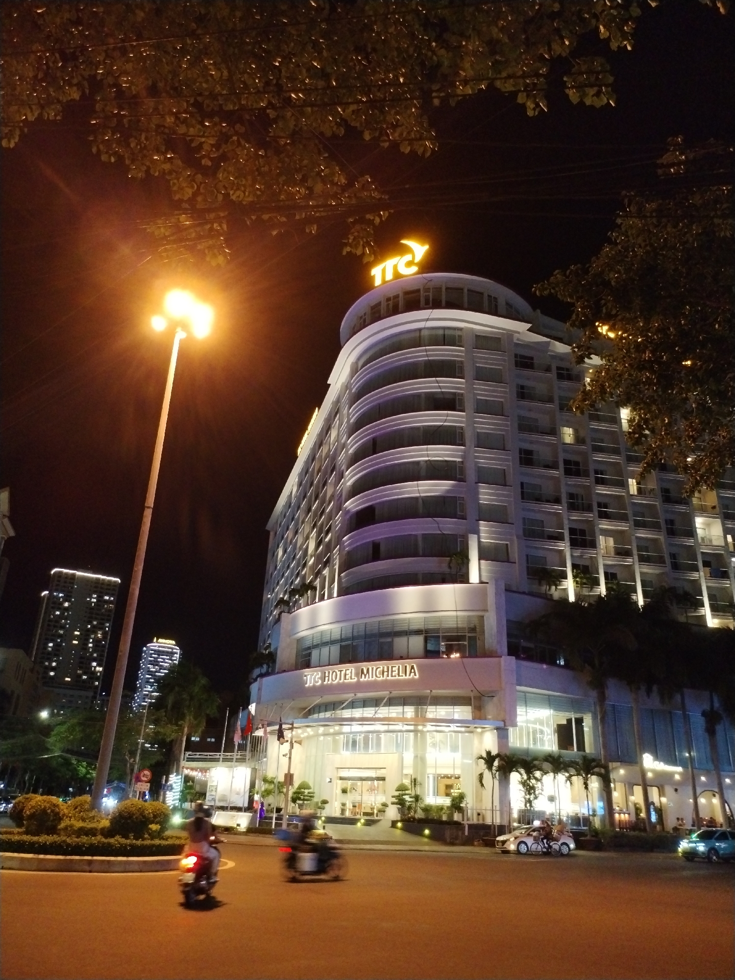 The photograph captures a night scene in Nha Trang Ward, Vietnam. Dominating the frame is the TTC Hotel Michelia, a modern, multi-story building with a curved facade, illuminated by both internal and external lighting, including a prominent 'TTC' logo on the top.
A bright street lamp casts a strong glare on the left side of the image, partially obscuring the night sky. The street is visible in the foreground, showing blurred figures on motorbikes suggesting movement. On the street level, there is a small garden.
Adjacent to the hotel, palm trees stand as silhouettes, along with other unidentified buildings in the background. The overall ambiance indicates an urban setting at night.