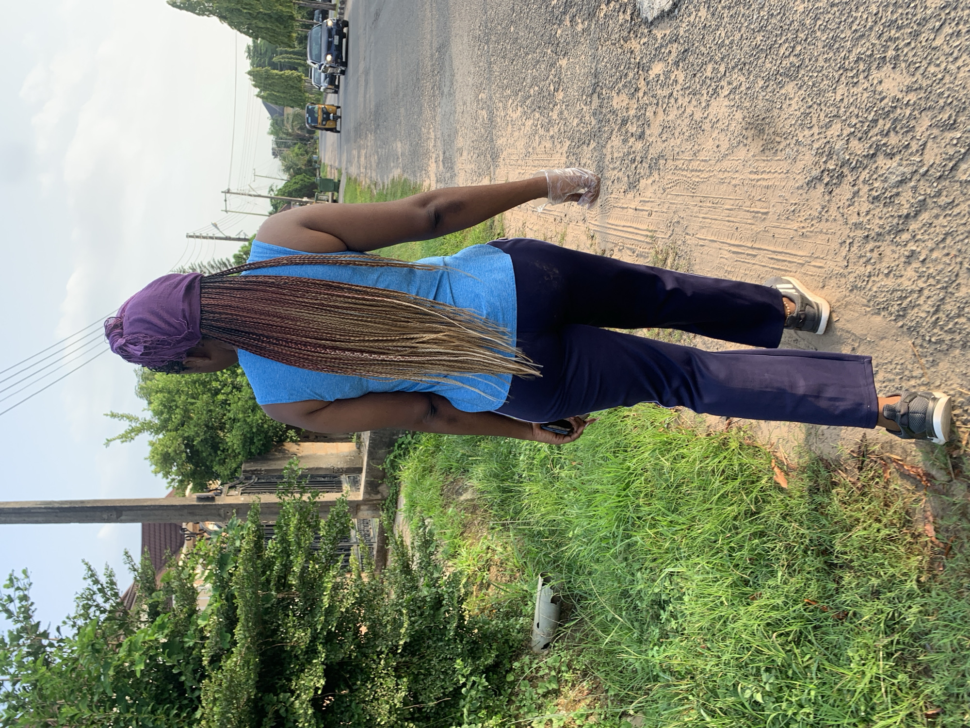 A person with long, braided hair is walking on a dusty path beside a road. The person is wearing a blue tank top and dark pants, with athletic shoes. They are also wearing a clear plastic glove on their right hand. The scene appears to be in a residential area of Abuja, Nigeria, with houses and trees visible in the background. There are cars and a three-wheeled vehicle on the road. The sky is partly cloudy.