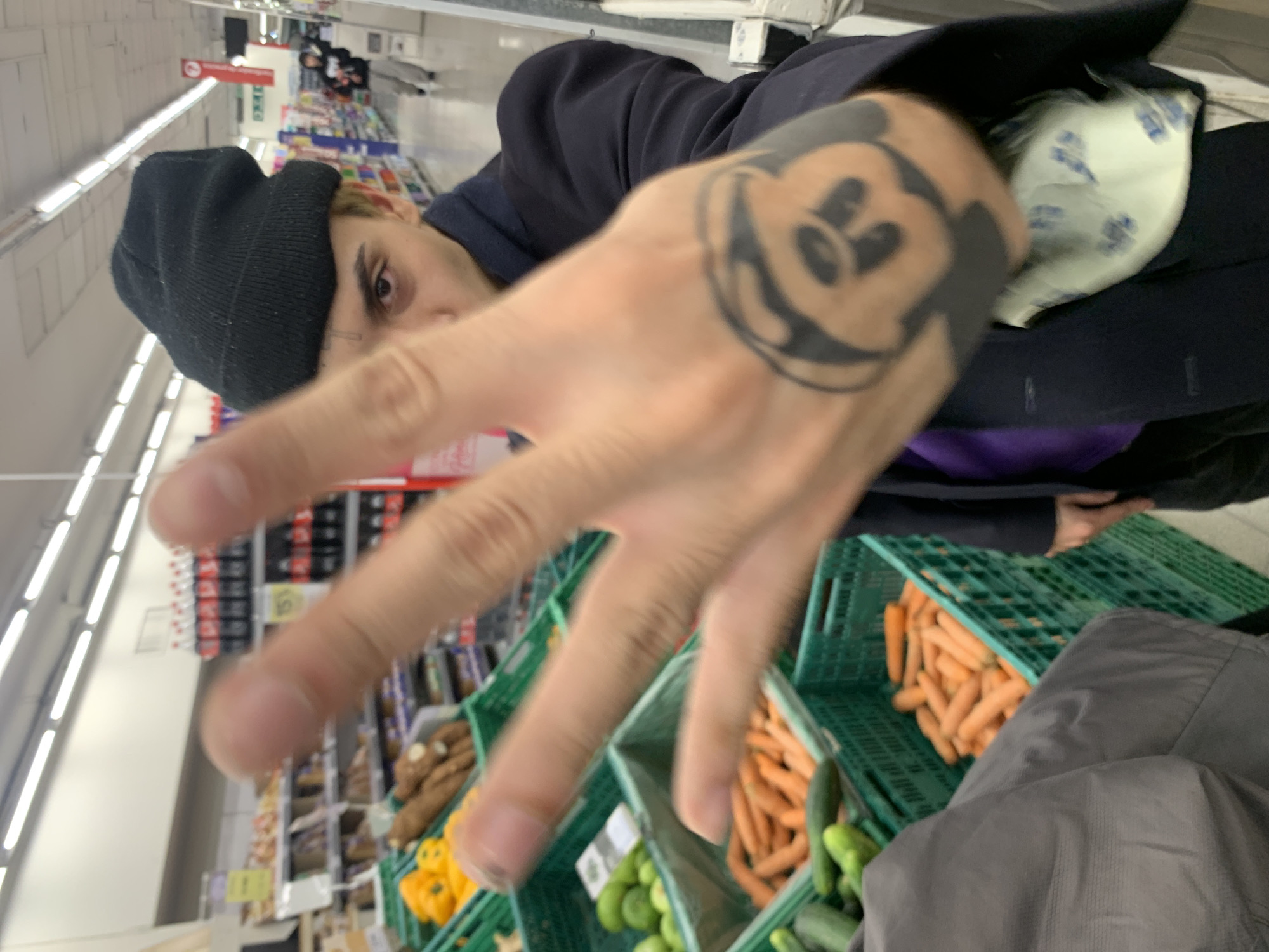 A person wearing a black beanie and dark coat stands in a grocery store aisle, holding up their hand with a Mickey Mouse tattoo. The person appears to be shopping for produce, with green crates containing cucumbers, carrots, and peppers visible in the foreground. Other grocery items are visible on shelves in the background. The location is a grocery store, but the specific location in Buenos Aires is not discernible from the image.