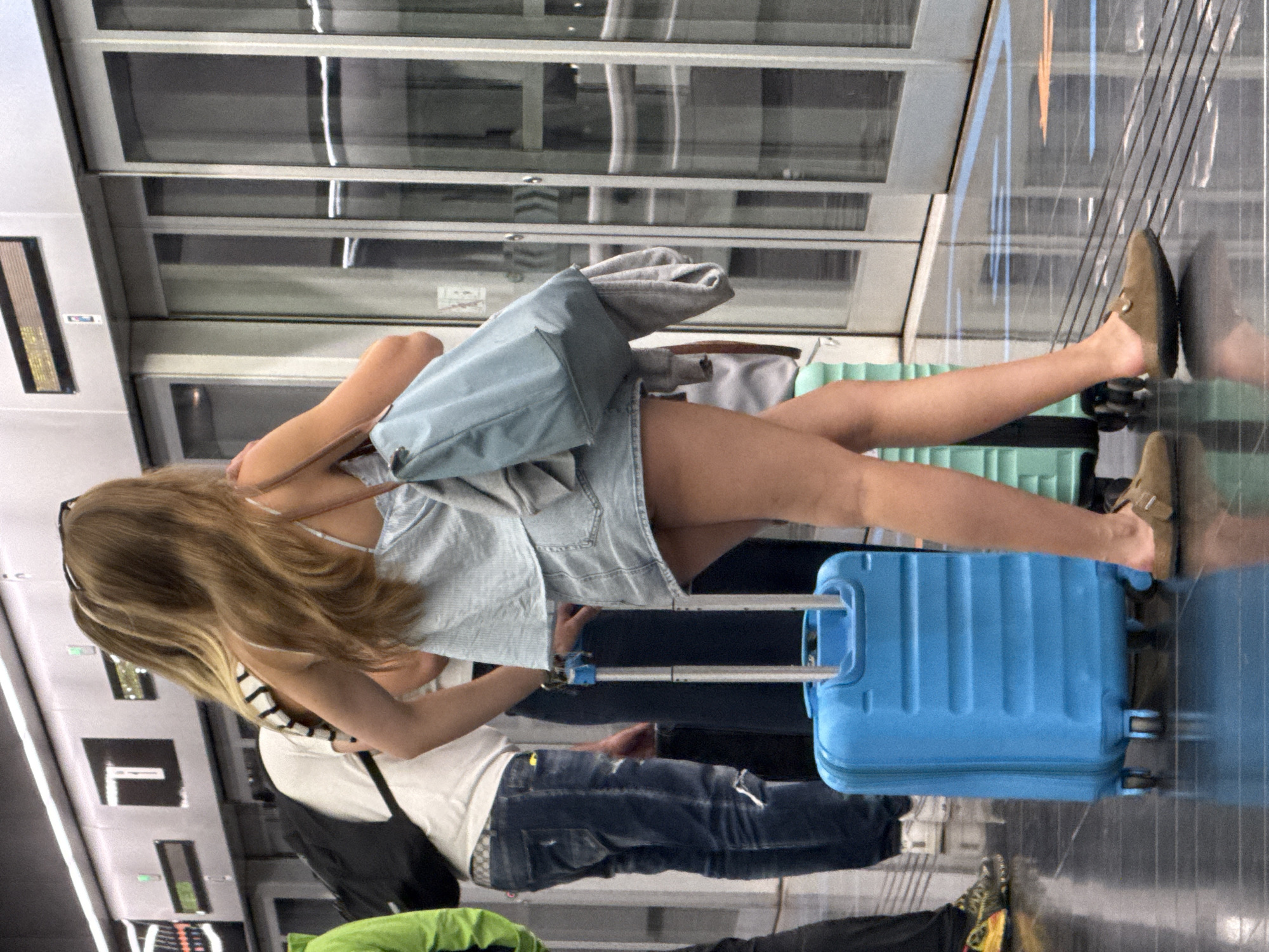 A group of people are standing on a modern, well-lit train platform. In the foreground, a woman with long blonde hair is facing away from the camera, wearing a light blue striped top and a denim skirt. She is holding a light blue tote bag over her shoulder, and a grey sweater is draped over her arm. Next to her, a bright blue suitcase with wheels stands on the polished floor, reflecting the lights of the platform. Behind her, another woman's back is visible, wearing a striped top. To the left, a person in a bright green jacket is seen from the back, carrying a black backpack. Another person in the background is wearing blue jeans and a white t-shirt. The train doors are visible behind the people, along with some signage on the wall. The overall environment suggests a transportation hub, likely within the city of El Prat de Llobregat, Spain, given the architecture and design elements commonly found in Spanish public transport stations.