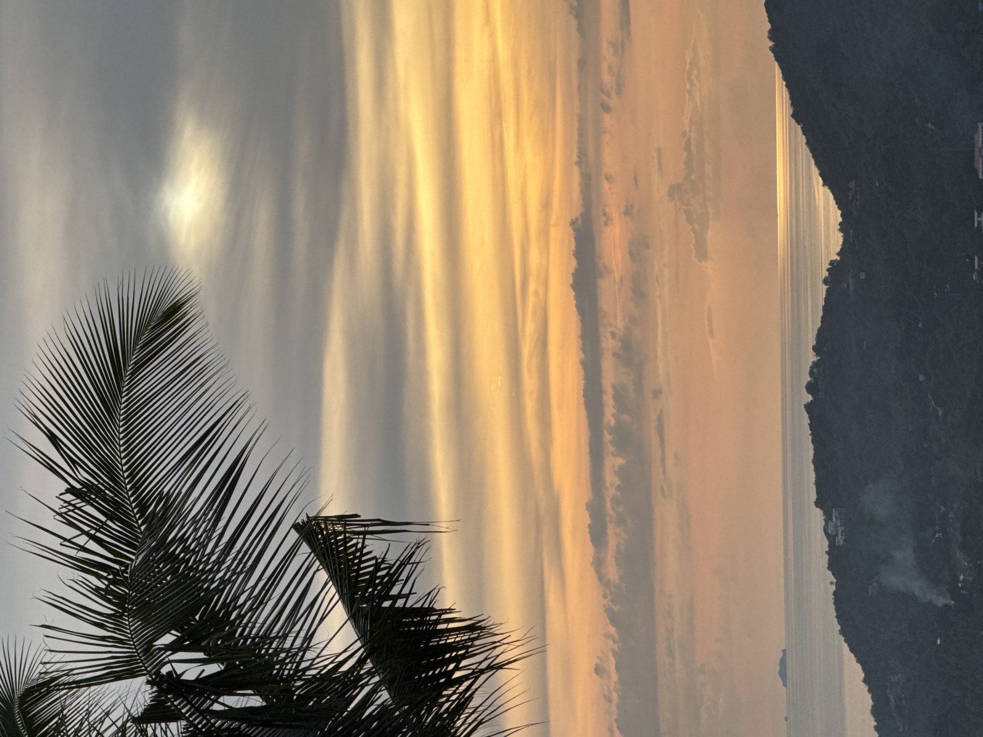 The image depicts a serene sunset scene over the ocean in Ban Tai Subdistrict, Thailand. In the foreground, a portion of a large palm frond is silhouetted against the sky on the left side of the frame. The sky itself is a gradient of colors, transitioning from a pale gray at the top to warm oranges and yellows near the horizon. Wispy clouds are scattered throughout, with a brighter patch of light indicating the setting sun peeking through.
Below the sky, the ocean stretches to the horizon, reflecting the golden hues of the sunset. In the distance, a small island or landmass is visible. The middle ground is dominated by lush, dark green hills covered in dense vegetation. Scattered across the slopes are hints of human habitation, with buildings and structures visible. A plume of smoke rises from one of the hillsides, suggesting a small fire or cooking activity. The overall impression is one of tranquility and natural beauty, characteristic of a tropical evening.
