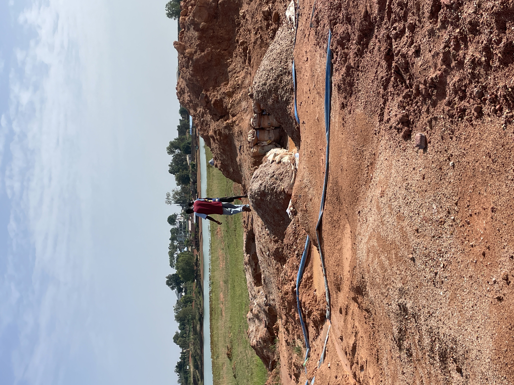 The image shows an outdoor scene near a body of water, likely in the city of Jos, Nigeria. The foreground features a steep, reddish-brown, earthen embankment. A blue pipe runs along its surface. Two individuals, wearing dark shirts and light-colored jeans, stand on a higher part of the embankment closer to the water. Behind them is a collection of stacked sacks. In the distance, across the body of water, are trees and buildings. The sky is pale blue.