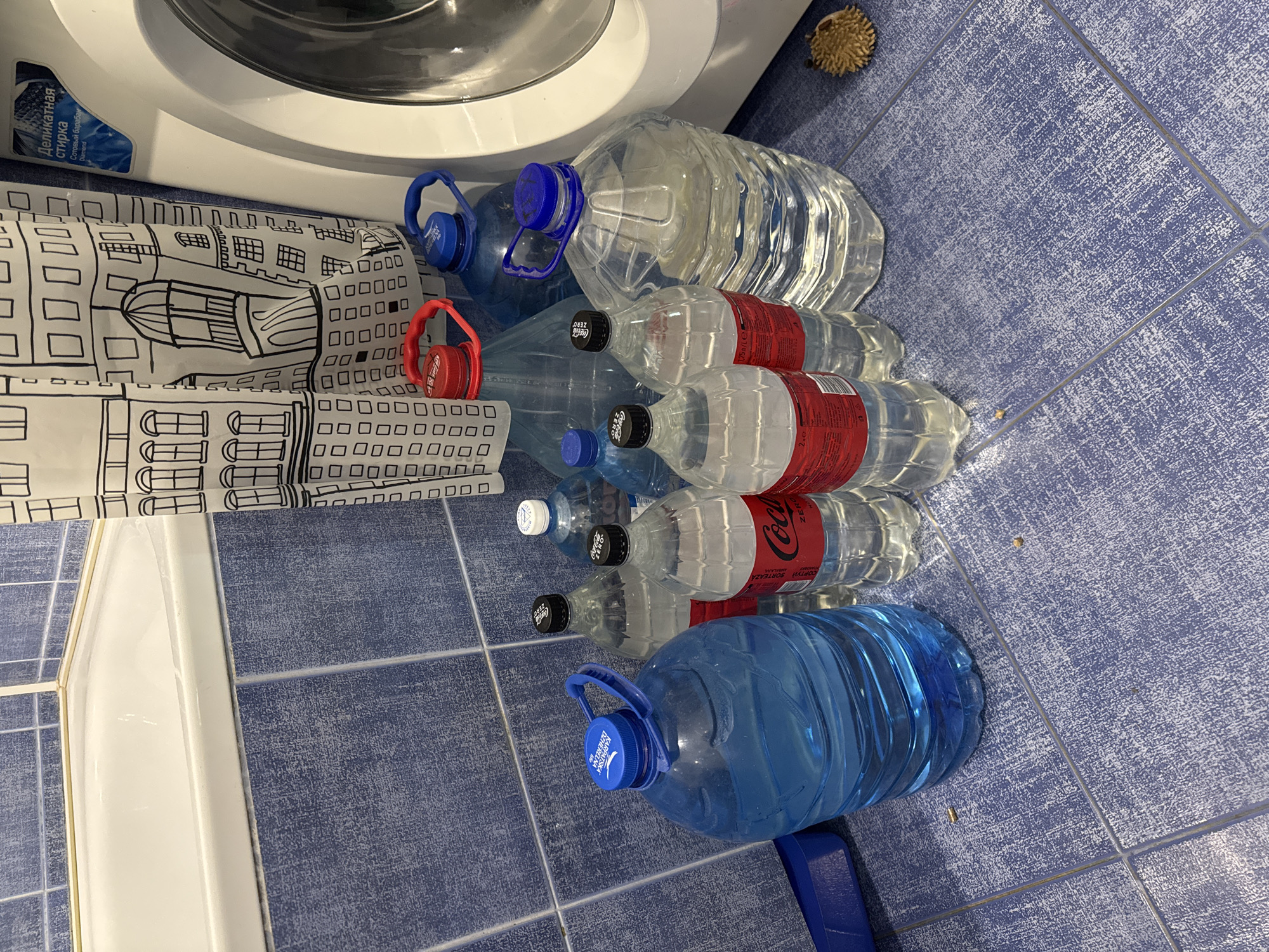 The image shows a collection of large plastic water bottles, some filled with clear liquid and others with blue liquid, arranged on a blue tiled floor. Some bottles have red or blue caps. In the background, a white bathtub with a blue tiled surround is visible. To the right of the bottles, a portion of a white washing machine is present, with a curtain featuring a black and white architectural pattern hanging beside it. A small, spiky object, possibly a decorative item, rests on the floor near the washing machine. The scene appears to be in a bathroom.