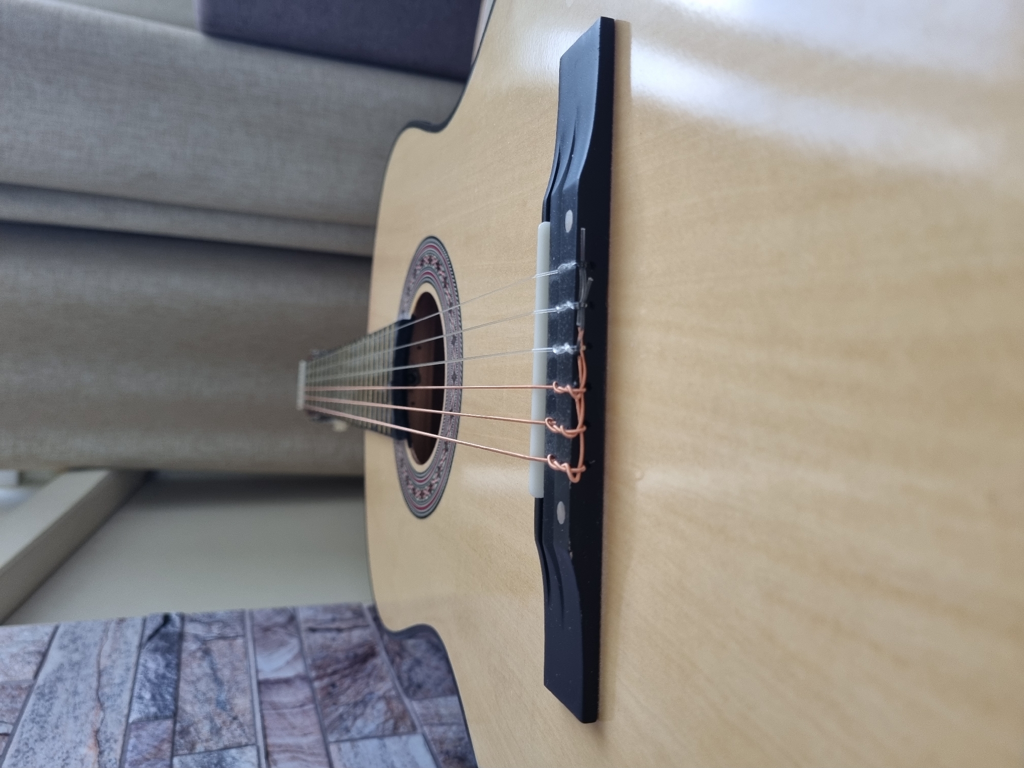 A close-up shot shows a light-colored acoustic guitar. The focus is on the guitar's bridge, which is black and holds the ends of the copper-colored strings. The guitar's body extends out of focus to the right, revealing a light wood grain. The soundhole with its decorative rosette is visible in the center. The background includes a neutral-colored upholstered item and a section of wall decorated with stone-like tile. No people are visible. There is no indication that the picture was taken in Sochi, Russia.