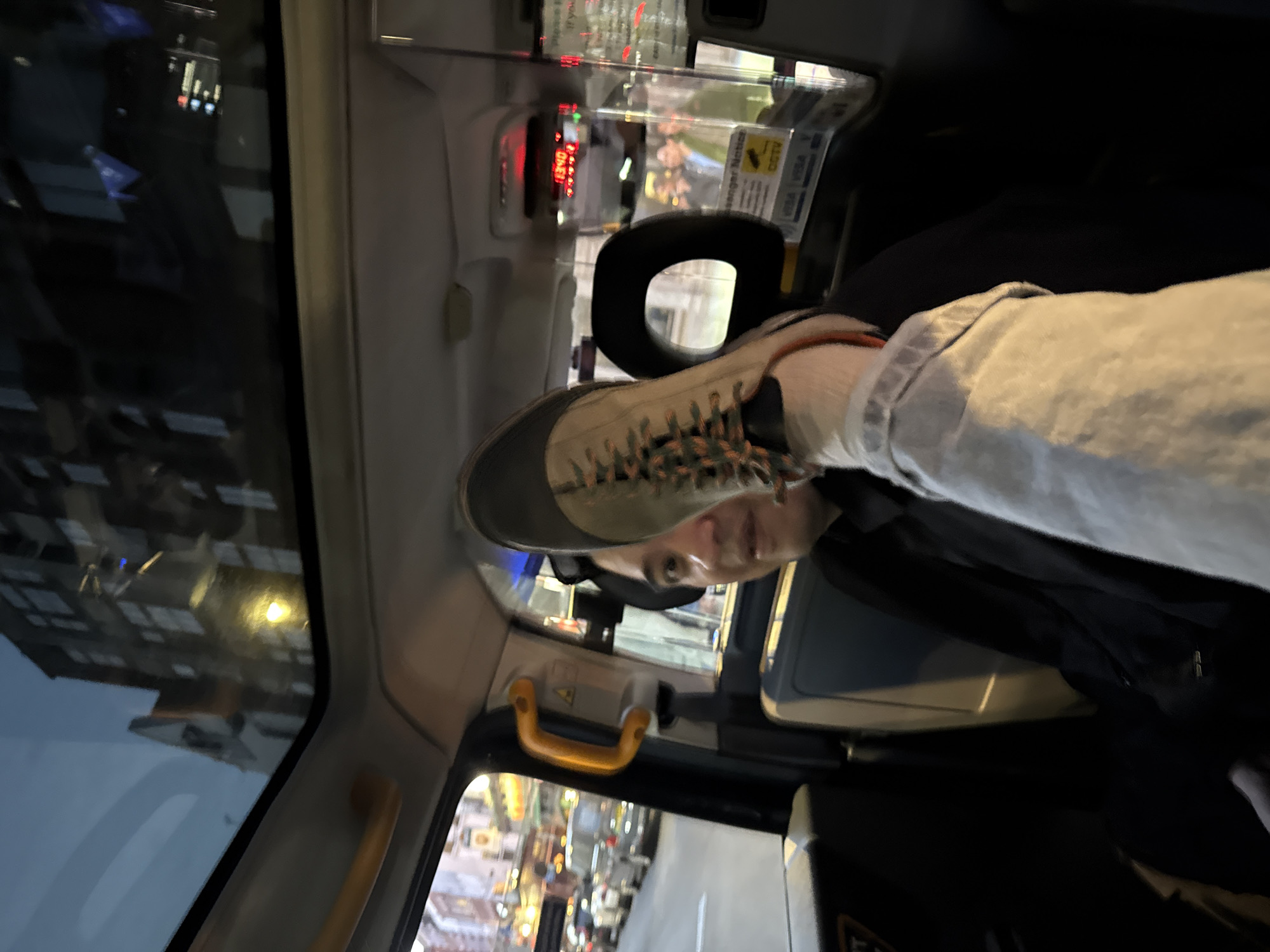The image shows the interior of a black London taxi, with a person's leg and foot, shod in a sneaker, resting on the seat headrest in front of them. The taxi's meter is visible, along with the typical plastic safety partition. Through the taxi window, a blurry view of a London street at night can be seen, with buildings and streetlights. The image appears to be taken by the passenger with the elevated foot.