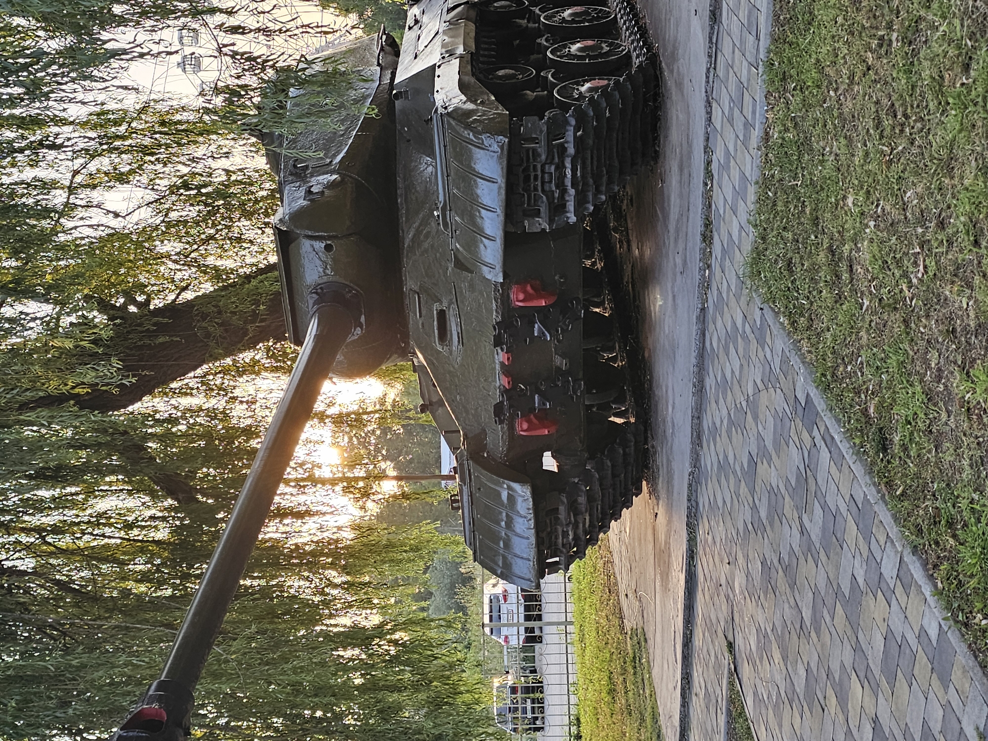 A dark green, tracked military tank is displayed on a paved walkway. The tank's long barrel extends to the left of the frame. The tank is positioned under the drooping branches of a large tree, with sunlight filtering through the leaves. In the background, a fence separates the display area from a road where two cars are parked. Beyond the road, a ferris wheel with several cabins is visible against a bright sky. The scene appears to be an outdoor monument or park. This image was taken in the city of Cherkessk, Russia.