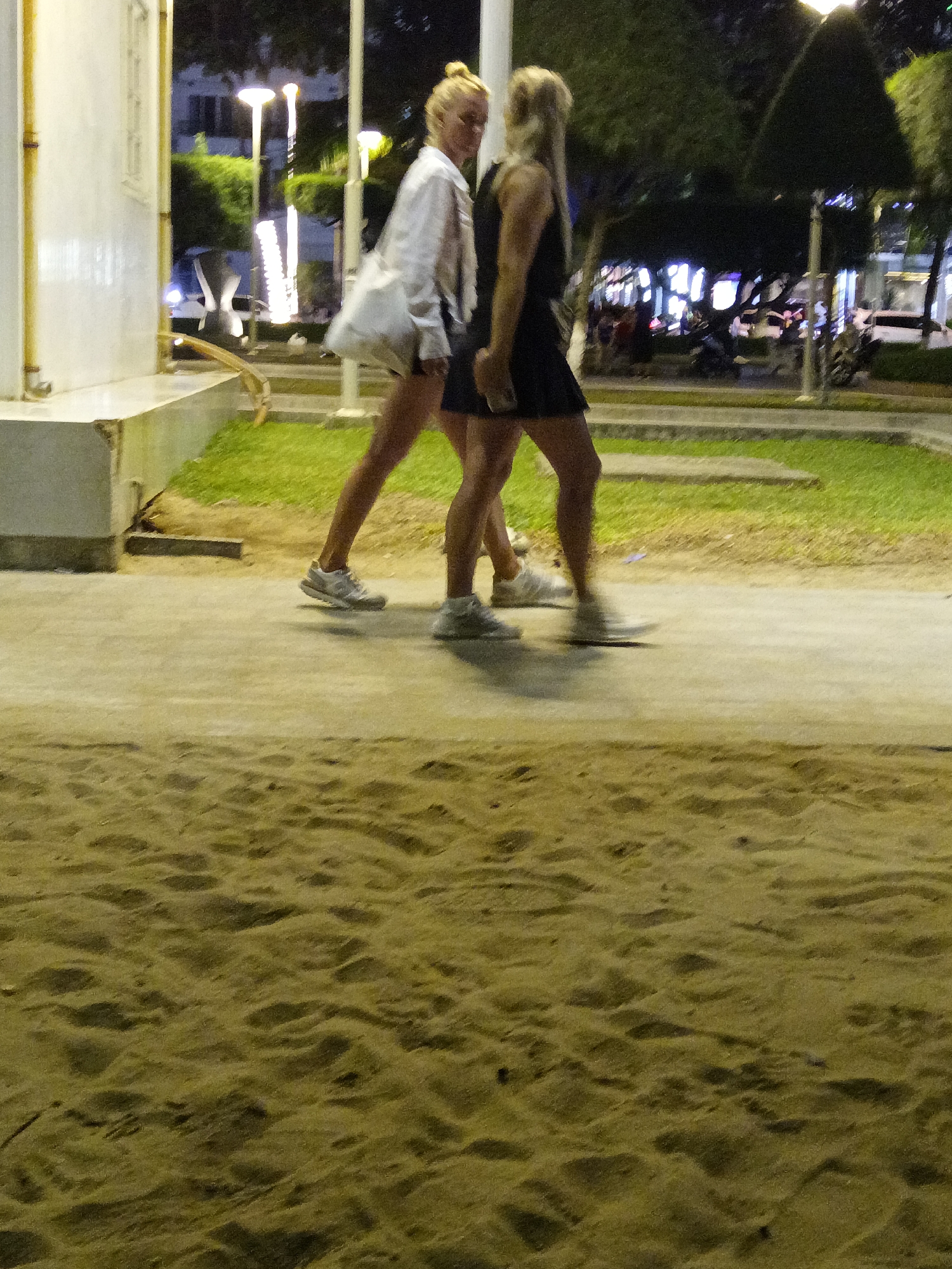 Two individuals, appearing to be women, are walking on a paved pathway next to a sandy area. The woman on the left is wearing a white shirt and shorts, carrying a white bag over her shoulder. She has blonde hair tied up in a bun and is wearing white sneakers. The woman on the right is wearing a dark-colored dress and dark sneakers, with blonde hair. They are walking in the same direction, away from the viewer. The background shows trees, streetlights, and distant buildings, suggesting an urban environment at night. The foreground consists of sand with visible footprints. The scene appears to be in Nha Trang, Vietnam, possibly along a beach promenade or park.
