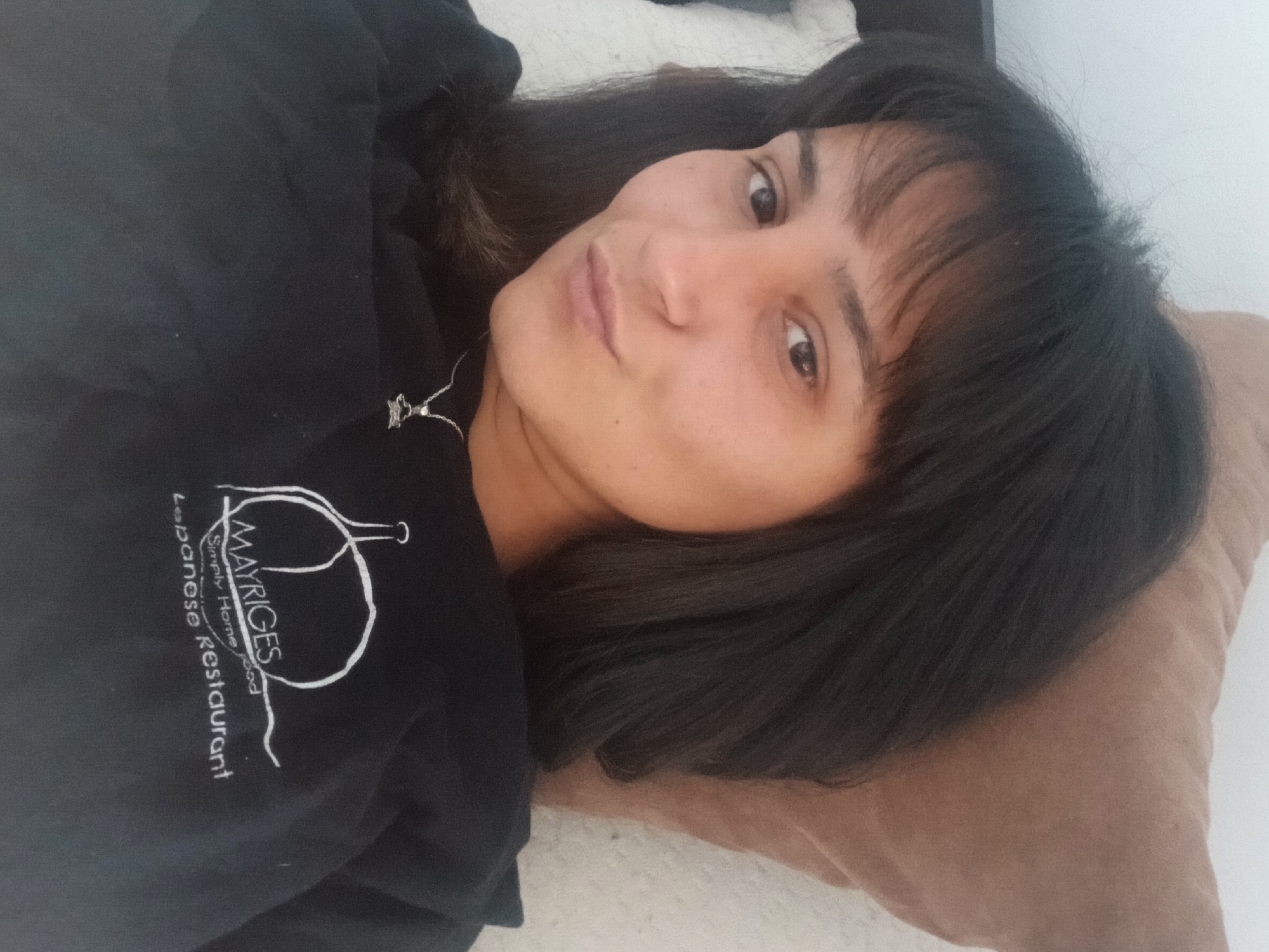 A close-up, slightly angled, eye-level selfie shows a young adult female with dark hair and bangs lying on a bed. She is looking directly at the camera with a neutral expression. She is wearing a black crew-neck sweatshirt with a white logo on the left side of the chest that reads "MAYRIGES Simply Home Food Lebanese Restaurant." She is wearing a silver necklace with a small pendant. The background consists of a beige pillow and a white wall. The lighting suggests an indoor setting during the day.
