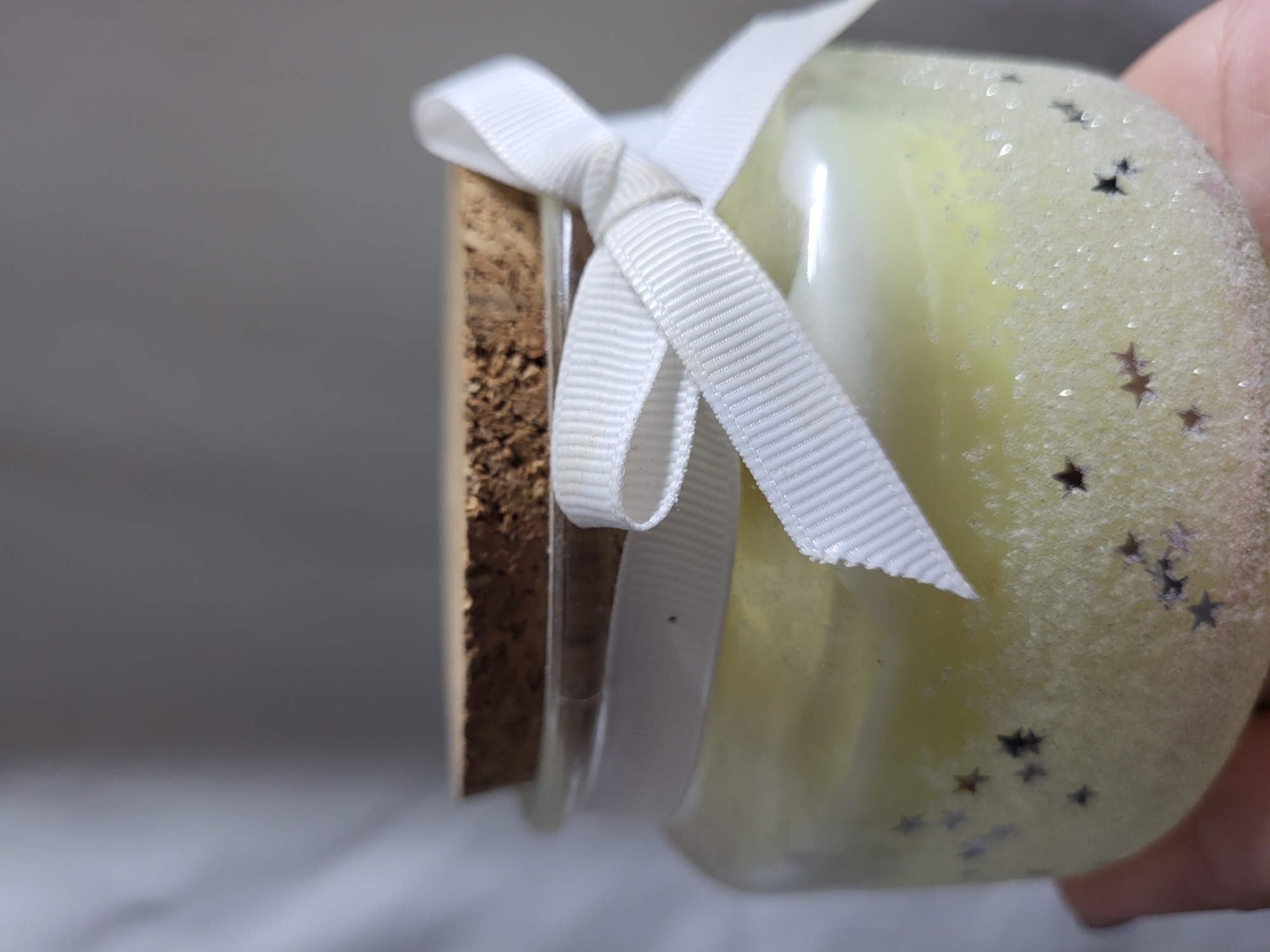 A close-up shot shows the top of a jar candle. The candle is pale yellow and contains small, dark star-shaped glitter pieces mixed into the wax. The jar has a light coating of what appears to be sand or fine glitter around the bottom portion. A white ribbon is tied in a bow around the neck of the jar, just below a cork stopper. The background is a soft, blurred gray and white fabric. The image appears to have been taken indoors, likely in a home environment in Horishni Plavni, Ukraine. There are no people or specific landmarks visible in the image.