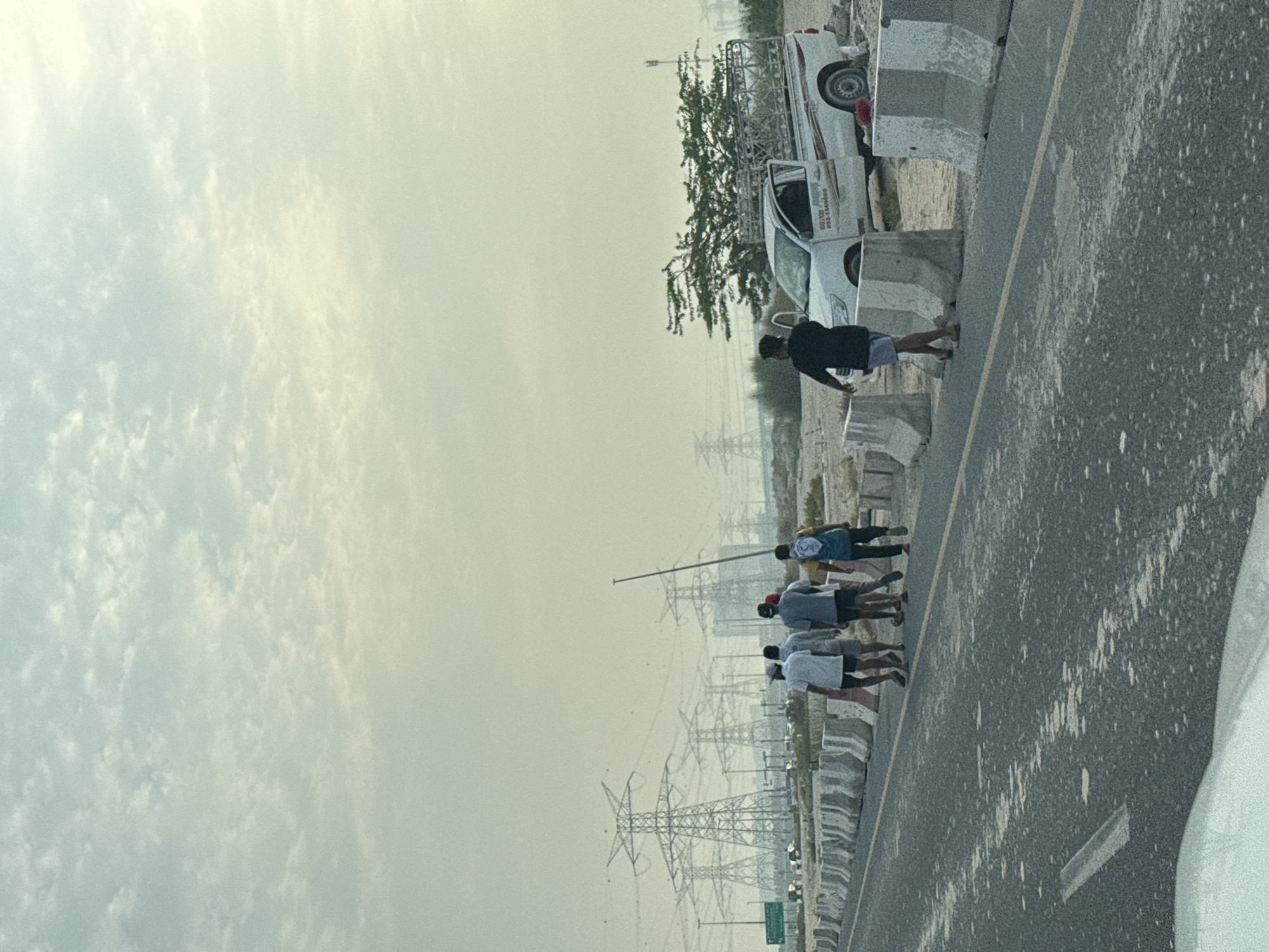 A group of five men walk along a paved road bordered by concrete barriers. In the background, several large electrical transmission towers are visible, suggesting an area with significant power infrastructure. A white pickup truck with its door open is parked on the sandy shoulder of the road, to the right of the walking men. The sky is overcast with a pale, diffused light. The environment appears to be a semi-arid or desert landscape on the outskirts of Dubai, United Arab Emirates, likely an industrial or infrastructure zone.