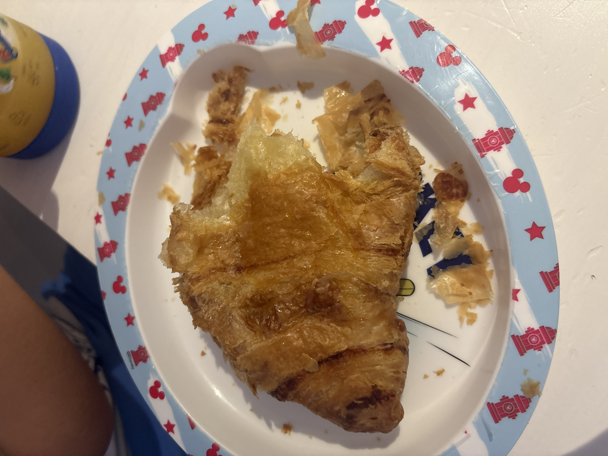 A close-up, overhead shot captures a half-eaten croissant on a white and light blue child's plate decorated with red Mickey Mouse heads and fire hydrants. The croissant, golden brown and flaky, has a large piece missing from its right side, with crumbs scattered around it on the plate. In the background, a blurred yellow and blue cup is visible. The scene takes place indoors, on a white surface, possibly a table. No specific location in Dubai, United Arab Emirates can be identified from the image.