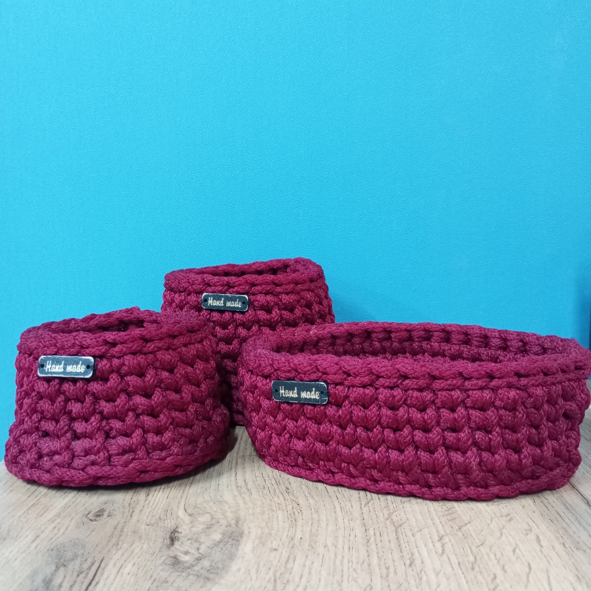 The image shows three handmade, deep-purple crocheted baskets on a light-wood surface against a textured turquoise wall. Each basket has a silver "Hand made" tag attached. The baskets are arranged in a staggered formation, with a rounder, taller basket in the center and a wider, more oval basket on the right. The wood surface appears to be a tabletop. The image provides no information to confirm the location is Pervomaysky, Russia.