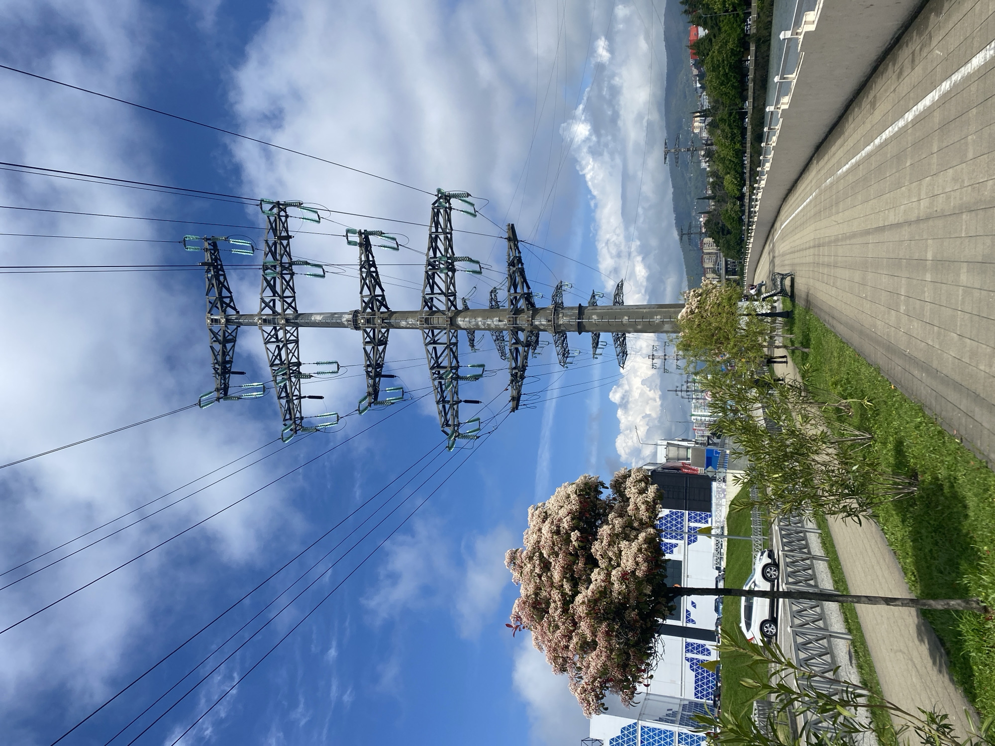 The image shows a view in Sochi, Russia, with a high-voltage power line pylon dominating the left side of the frame against a blue sky with scattered clouds. The pylon is metallic and supports numerous glass insulators. Power lines extend diagonally across the sky.
To the right, a paved walkway or road runs along a concrete barrier. Grass grows along the side of the road. A tree with light pink blossoms is situated next to the walkway. In the background, buildings and trees are visible on a hillside. A white vehicle can be seen parked nearby. Two people are sitting on a bench on the side of the road.