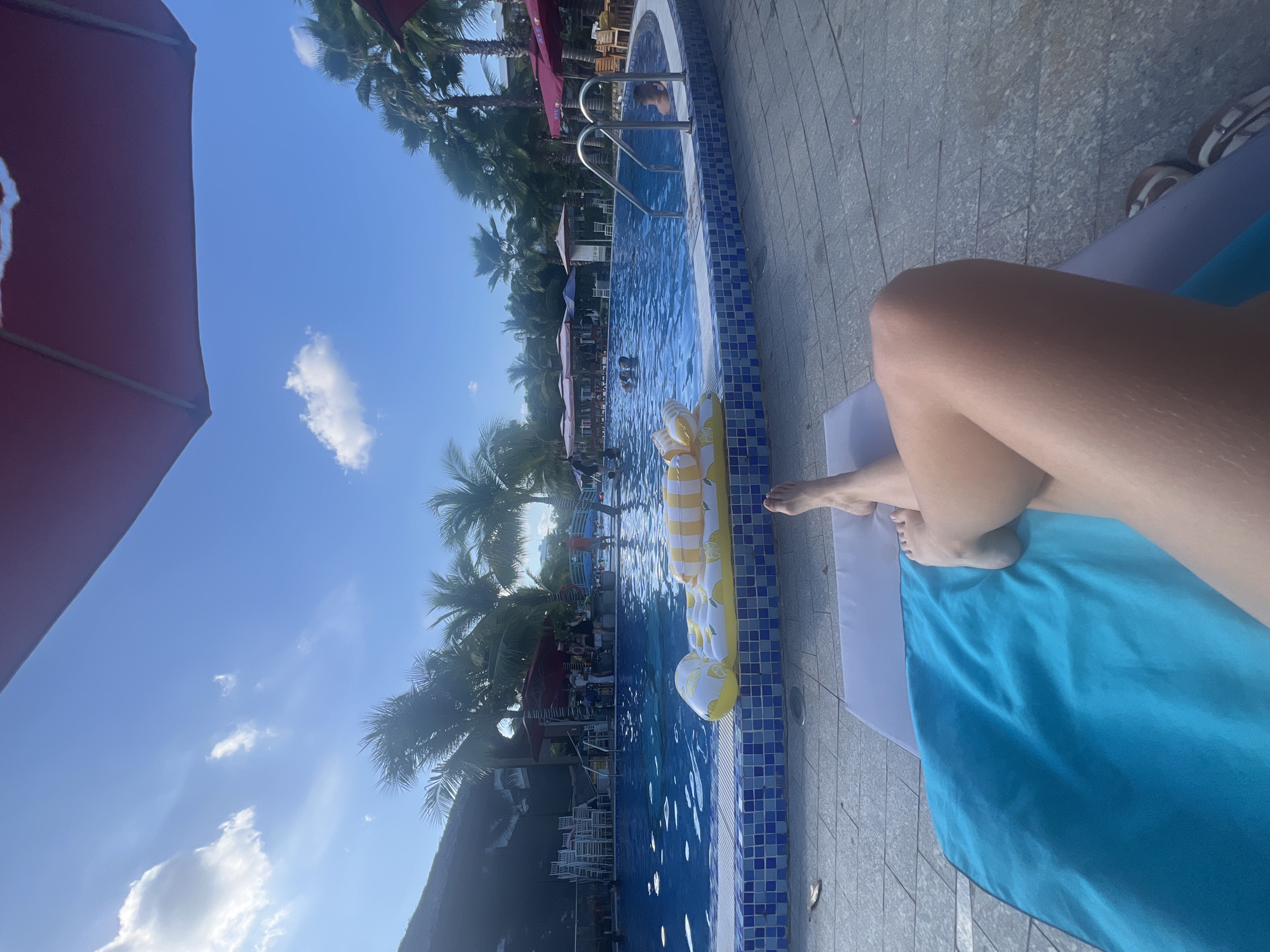 The image shows a poolside scene in Nam Nha Trang Ward, Vietnam. A person is lying on a blue towel on a lounge chair next to a swimming pool. The pool has a yellow and white inflatable raft shaped like a lemon floating in it. The pool is surrounded by palm trees and umbrellas. The sky is blue with scattered clouds. Other people are in the pool and on the pool deck in the background.