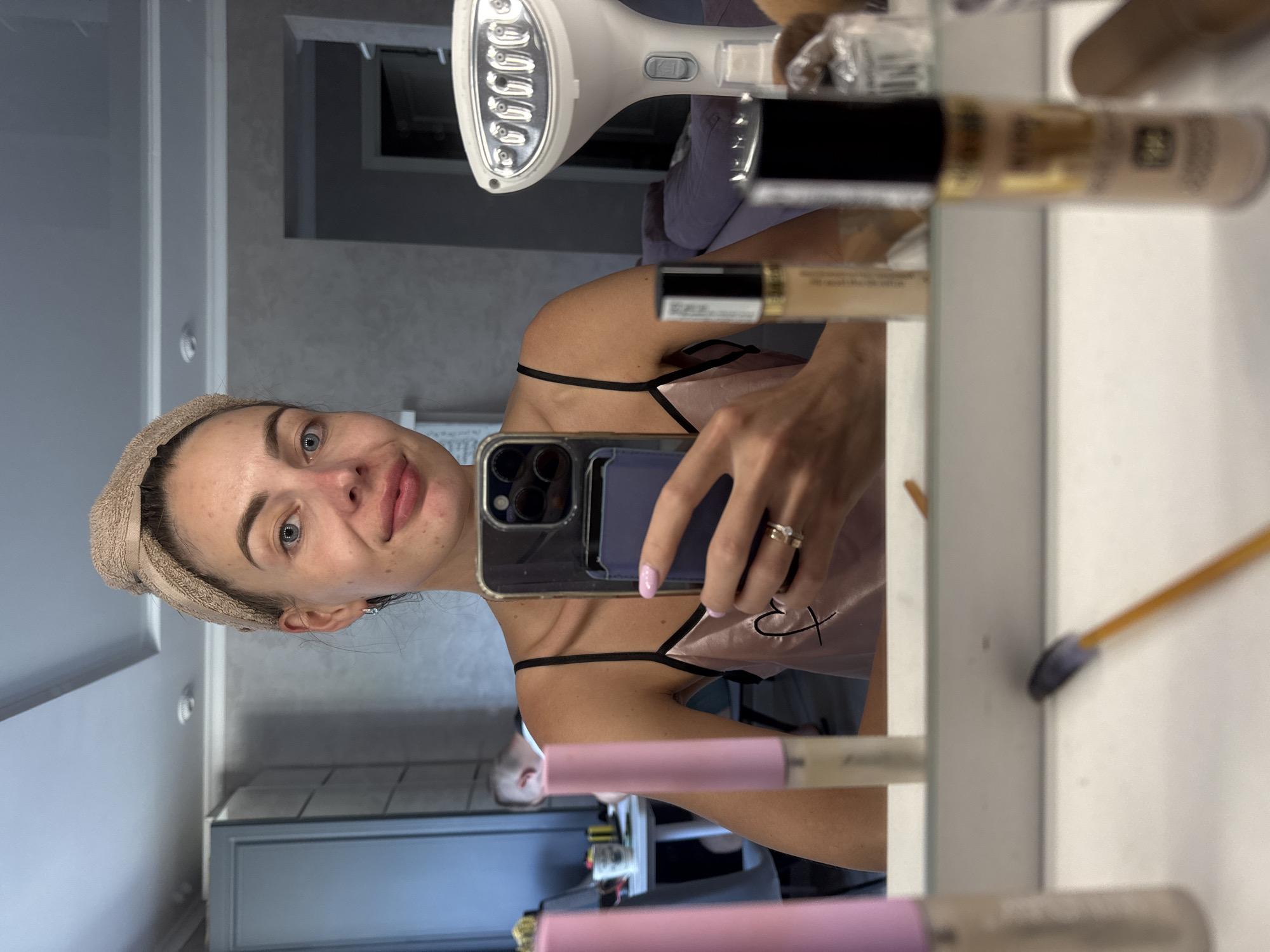 A woman is taking a selfie in a mirror. She has a towel wrapped around her head and is wearing a pink camisole. Her face is bare, and she has a slight smile. In the mirror, we can see some makeup products on a vanity, as well as a garment steamer. In the background, a man is visible sitting at a table. The scene appears to be in a room, possibly a dressing room or bedroom. There are no specific indicators of the location being in Sochi, Russia.