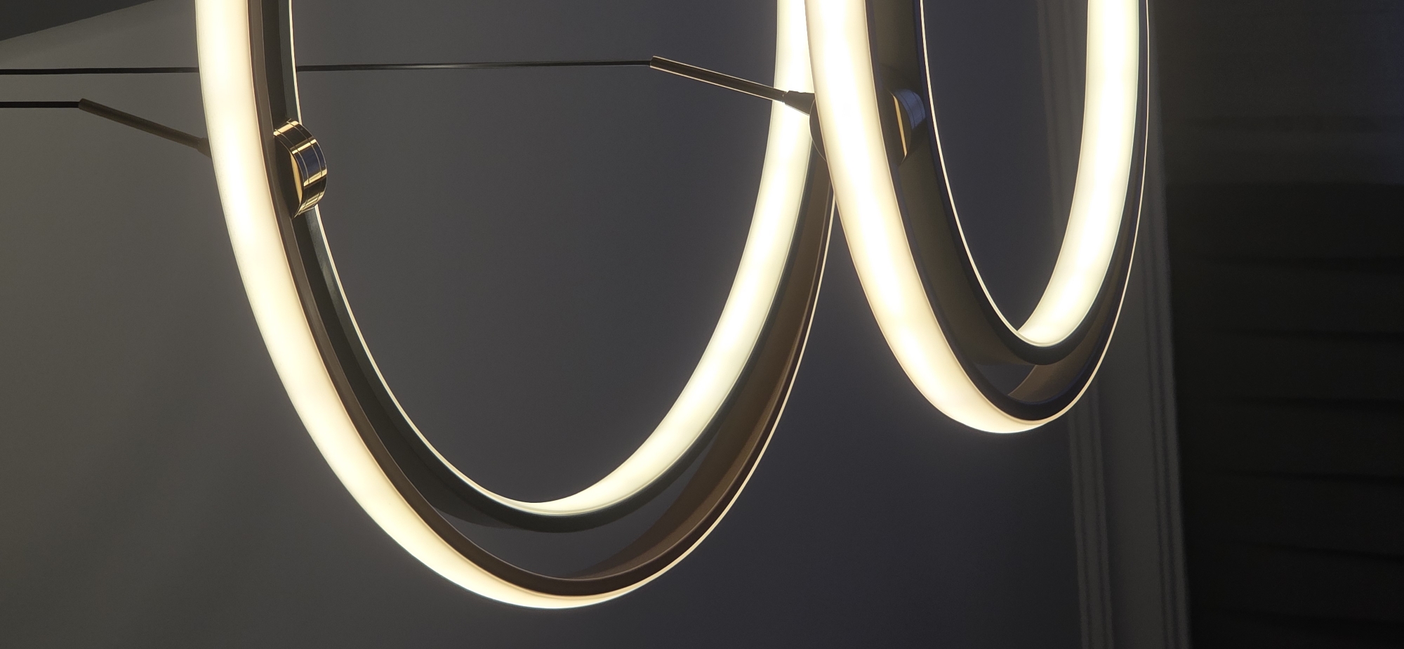 The image shows a close-up view of a modern, circular pendant light fixture with two illuminated rings. The fixture is suspended from the ceiling by thin black wires. The rings appear to be made of metal with a dark finish, and they have a warm, white LED light source integrated into their inner edges. The background is a dark, solid color, likely a ceiling or wall, with subtle shadows and highlights indicating the lighting from the fixture. There are no people, animals, or any other objects visible in the frame. The scene is indoors, and the overall impression is one of minimalist and contemporary interior design. There is no visible indication of the city of Sumgayit, Azerbaijan, or any specific location within it.