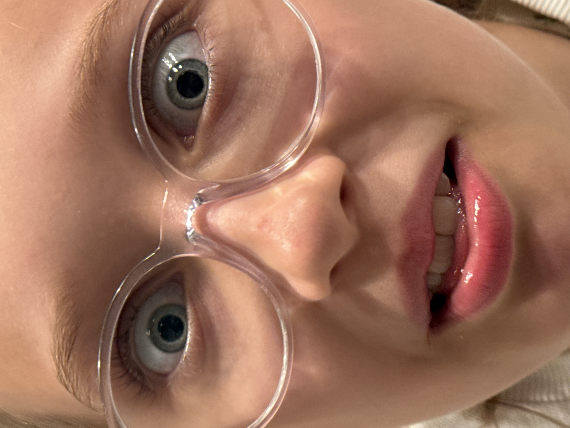 This is a close-up, vertical, overhead shot of a young person's face, oriented sideways, with their mouth and eyes visible. They are wearing clear-rimmed glasses, with the lenses framing their eyes. The person's skin appears fair. Their eyes are open, with the irises being a light gray-blue color and the pupils dilated. Their eyebrows are light brown and feathered. The mouth is open, revealing teeth, and the lips are glossy with a pink hue. The background is mostly out of focus but suggests a textured surface, possibly clothing, with a striped pattern in a light color. The lighting appears to be indoor ambient light, possibly from a ceiling fixture, creating some shine on the skin and lips. There is no visible text. The scene is a close-up portrait of a child, with no discernible setting cues beyond what can be inferred from the clothing. The emotions are difficult to ascertain due to the extreme close-up and static nature of the image, but the open mouth and wide eyes might suggest surprise, curiosity, or a playful expression. The image was likely taken in Riga, Latvia, as per the location context.