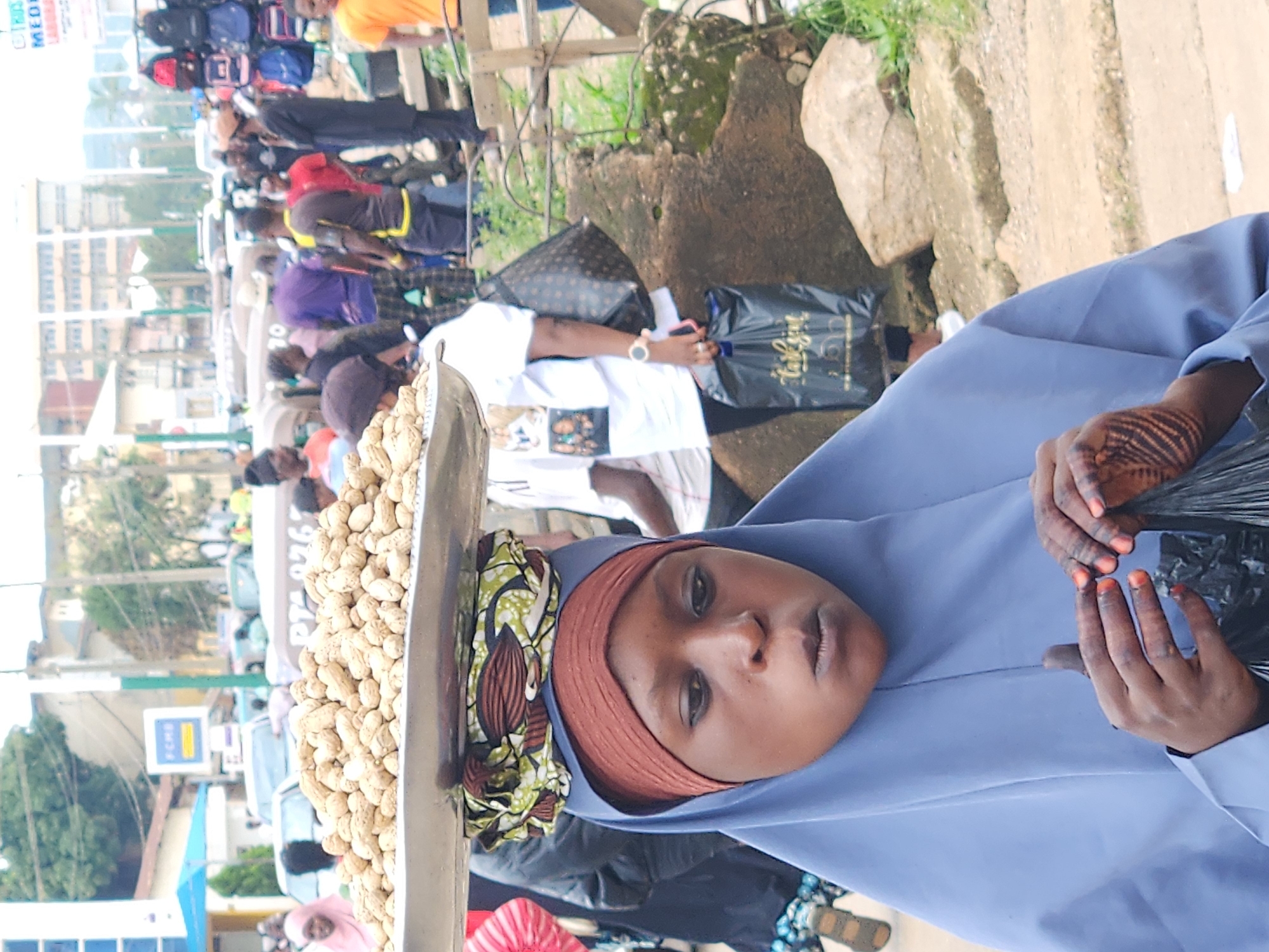 The image depicts an outdoor marketplace scene in an urban environment. In the foreground, a young woman wearing a blue hijab and a reddish-brown headscarf is the primary subject. She is carrying a large metal tray filled with peanuts balanced on her head, which is cushioned by a colorful patterned cloth. Her hands, adorned with henna, are holding a black plastic bag.
Behind her, other individuals are visible, some walking or standing, engaged in market activities. A variety of goods are displayed in the background, including what appears to be bags or backpacks on a rack to the right. The setting suggests a busy street or market with buildings and vehicles in the distance. The overall atmosphere is that of a bustling community gathering.