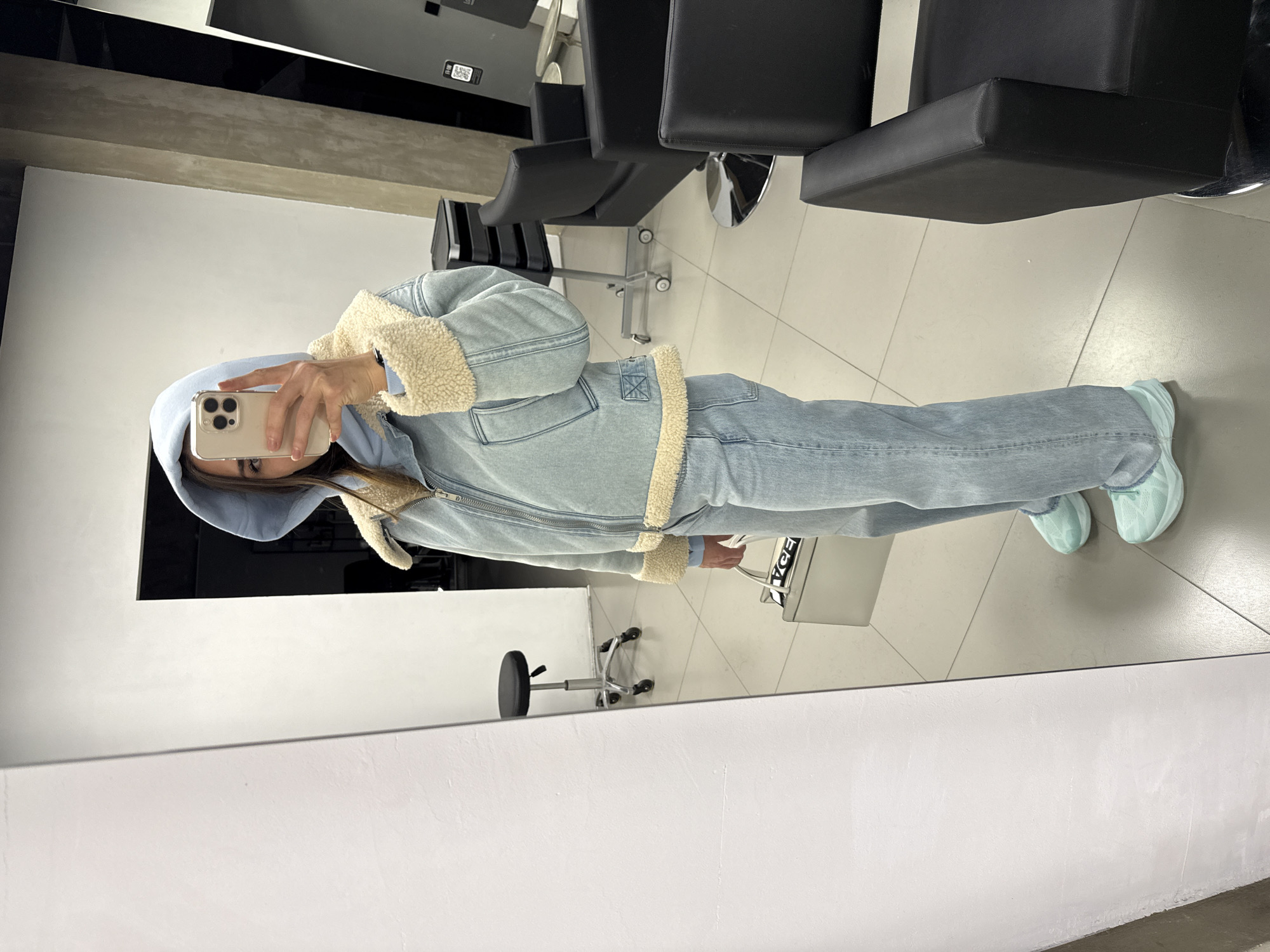 A person is visible in the reflection of a mirror in what seems to be a hair salon in Saint Petersburg, Russia. The person is holding a smartphone to take a photo of themself. They are wearing a light blue hooded sweatshirt and a denim jacket with a sherpa lining. They also have on light blue denim jeans and matching light blue sneakers. Behind them are several black salon chairs and a salon styling cart. The floor is covered in large, square, light-colored tiles. The reflection includes the salon furniture, walls, and the floor.