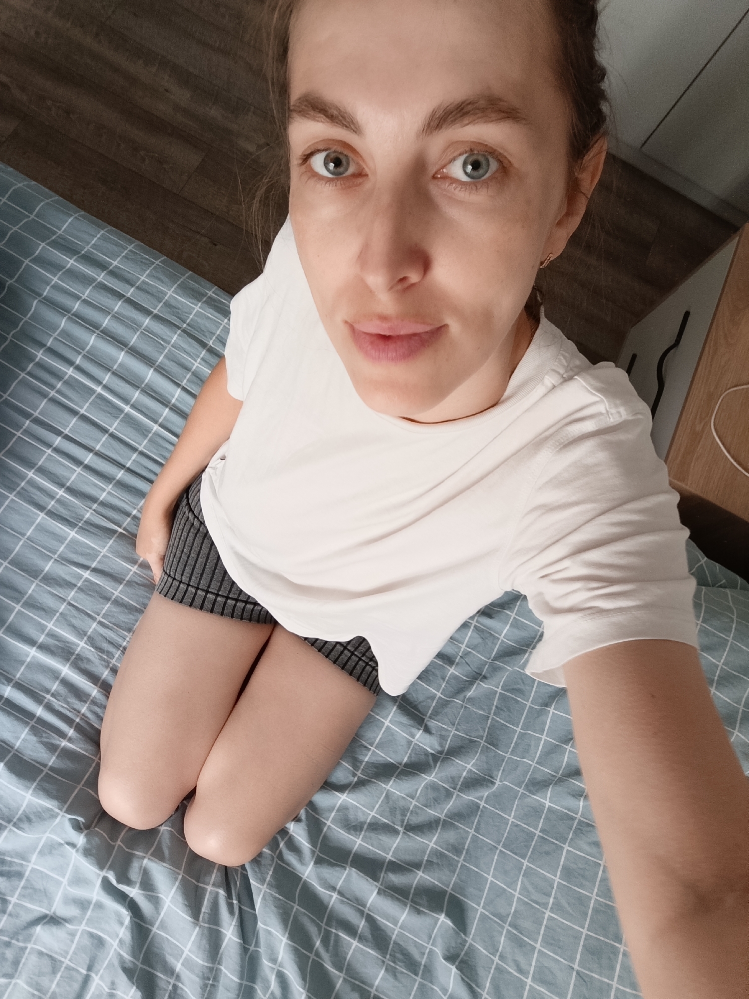 A woman is kneeling on a bed. She has light skin, blue eyes, and brown hair pulled back. She is wearing a white t-shirt and grey and black striped shorts. The bed has a blue and white checkered comforter. To the left of the bed is a wooden floor. To the right of the bed is a wooden nightstand with a white cord resting on it. The scene appears to be indoors in a bedroom.