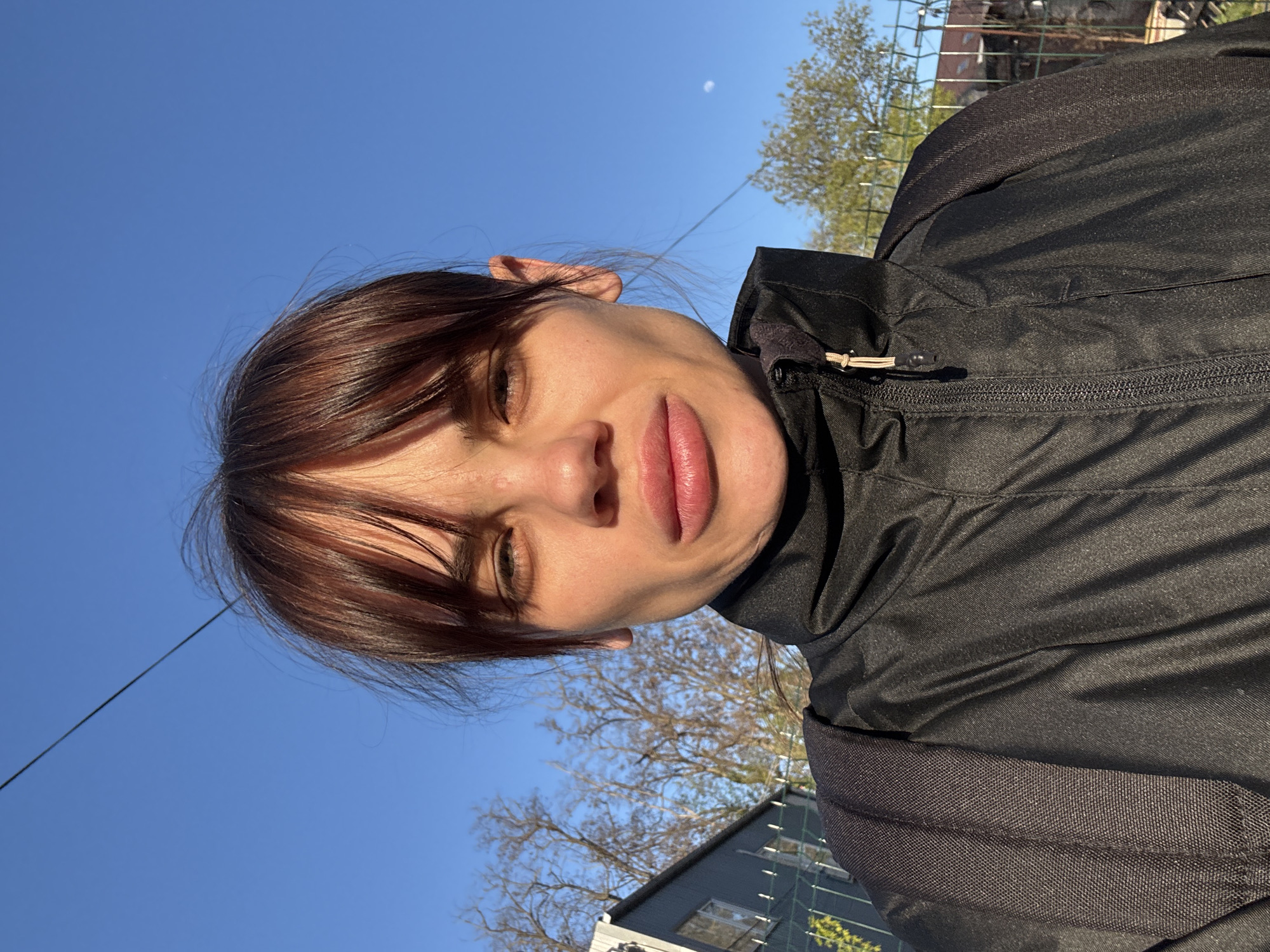 A close-up outdoor portrait, slightly tilted, features an individual with medium-dark brown hair and bangs, looking upwards under a clear blue sky. The subject, likely female, has fair skin with a neutral expression, and her eyes are partially closed or squinting, possibly due to bright sunlight. Full lips are prominent. She is wearing a black, high-collared jacket, which appears to be made of a synthetic material. Details of the jacket include a visible zipper and a small beige/tan cord with a toggle near the right shoulder, suggesting a functional outdoor garment or backpack strap.
The background is dominated by a vibrant, cloudless blue sky, indicating sunny weather. A faint, crescent moon is visible in the upper right sky, suggesting the photograph was taken during daylight hours when the moon is still present, possibly early morning or late afternoon. Sparse, leafless trees are visible against the sky, hinting at late winter or early spring. A thin black wire or cable stretches across the sky, passing near the subject's head. In the lower right background, a dark-colored building or structure with some visible windows or scaffolding can be seen, partially obscured by the trees and the jacket. A wire fence is also visible behind the subject's right shoulder.
The scene captures a casual moment outdoors, likely in an urban or suburban environment within Rostov-on-Don, Russia. The activity is a simple portrait or selfie under bright, clear weather conditions. There is no visible text in the image.