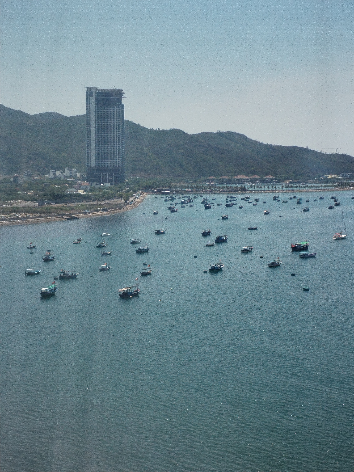 The image captures a wide view of the bay in Bac Nha Trang Ward, Vietnam, with a focus on the numerous fishing boats scattered across the water. The scene is set against a backdrop of rolling hills and a partially constructed, modern high-rise building that stands out prominently on the shoreline. The water is a calm turquoise-blue.